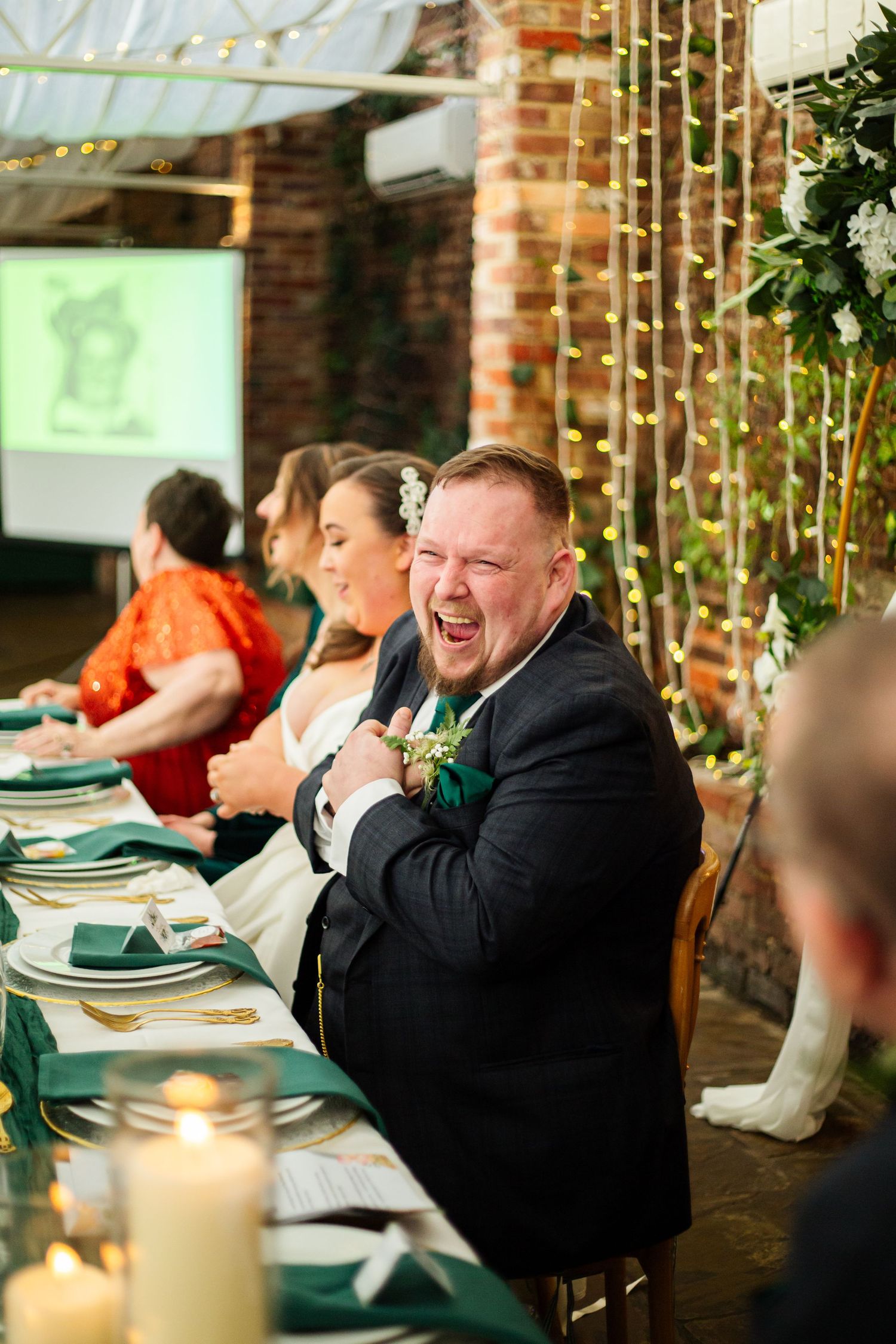 Wedding guests laugh and smile during dinner reception at Northbrook Park venue with twinkling lights and greenery decor.