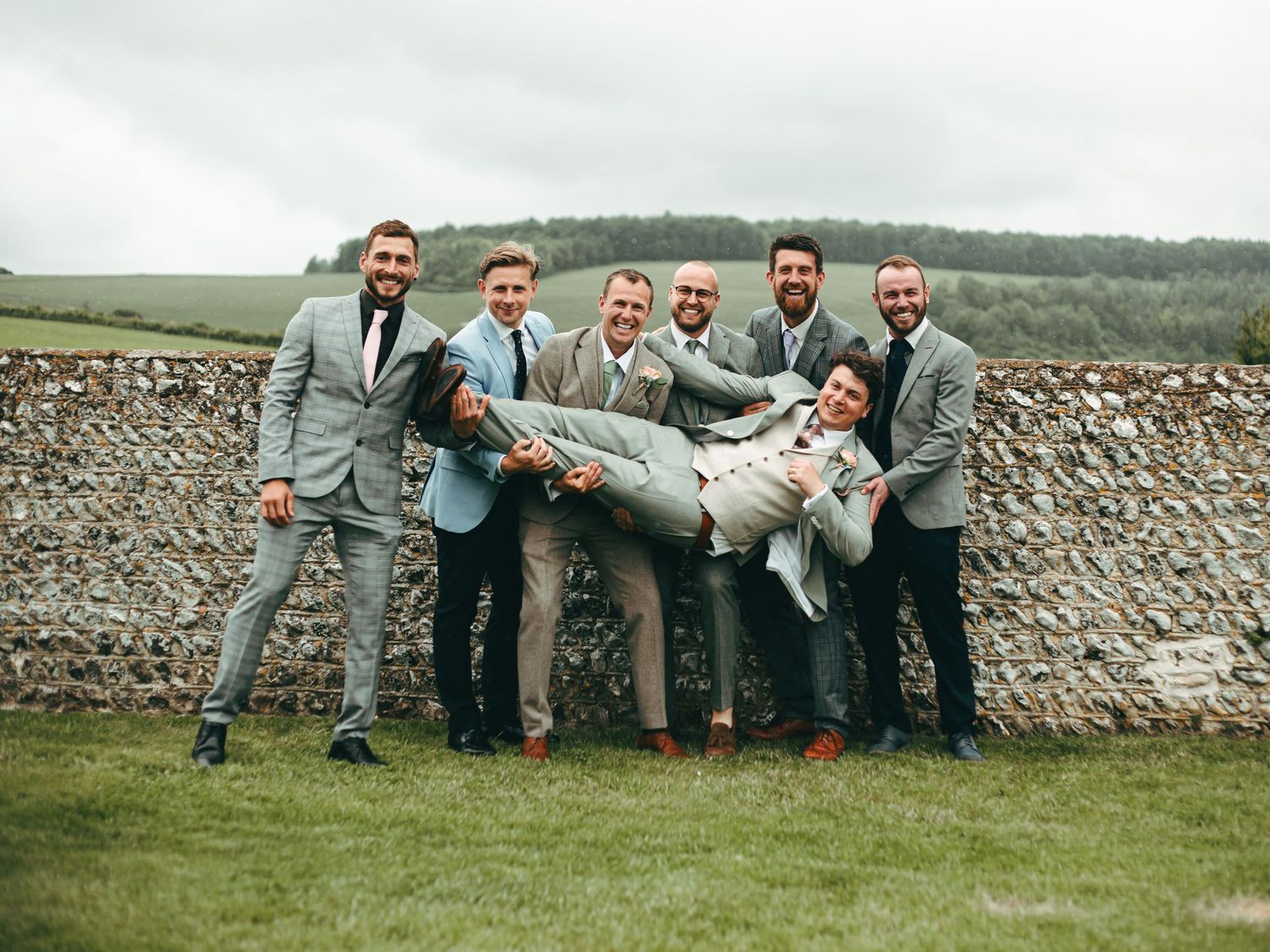 Groomsmen in matching gray suits playfully lift someone during outdoor wedding photo session by stone wall at Southstoke Barns near Arundel.