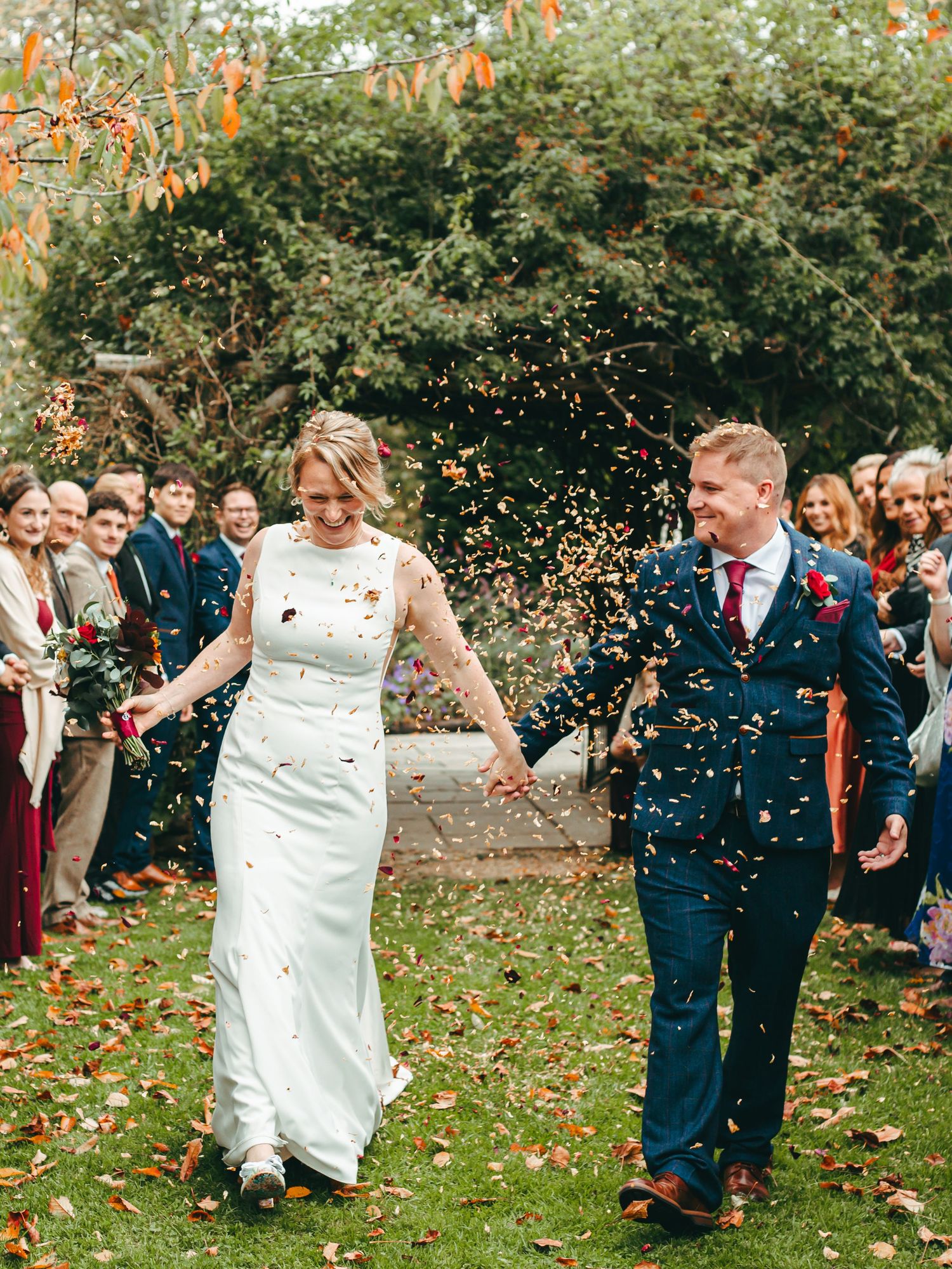 Wedding couple walks down garden path while guests throw confetti during autumn celebration with orange flowers overhead at Rumbold Farm