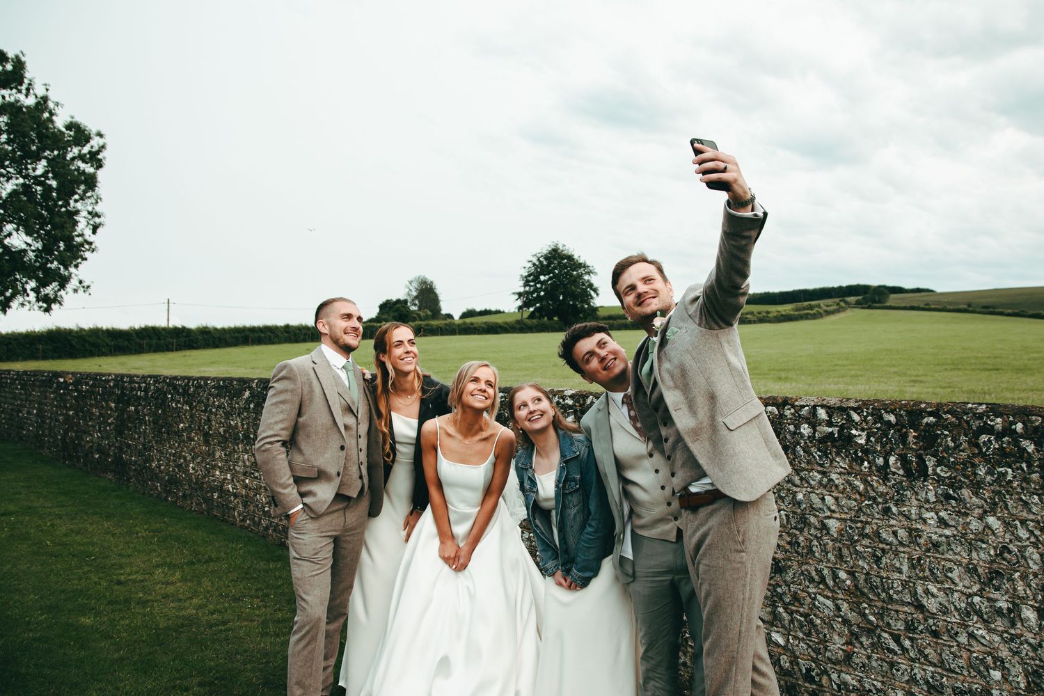 Wedding party poses playfully for photos in front of a rustic stone wall on a sunny day in the countryside at Southstoke Barn near Arundel
