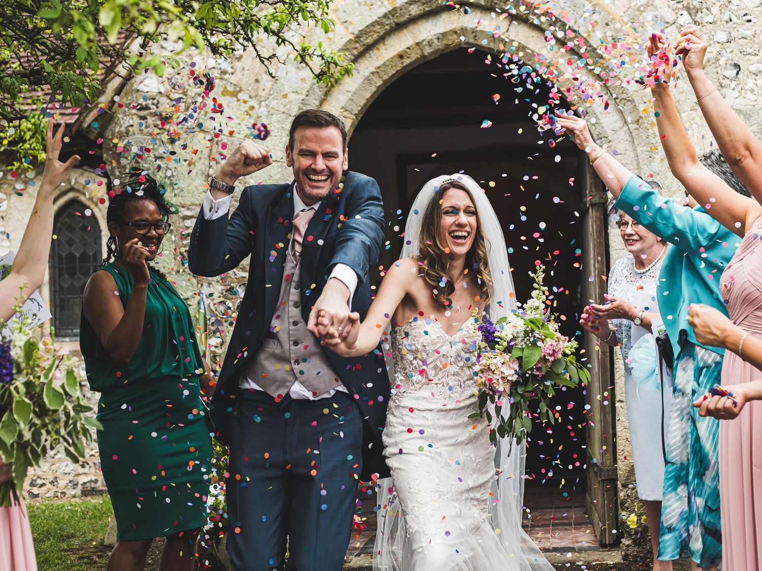 Wedding guests throw colorful confetti over newlyweds outside a stone church entrance during joyful celebration at Burpham Church
