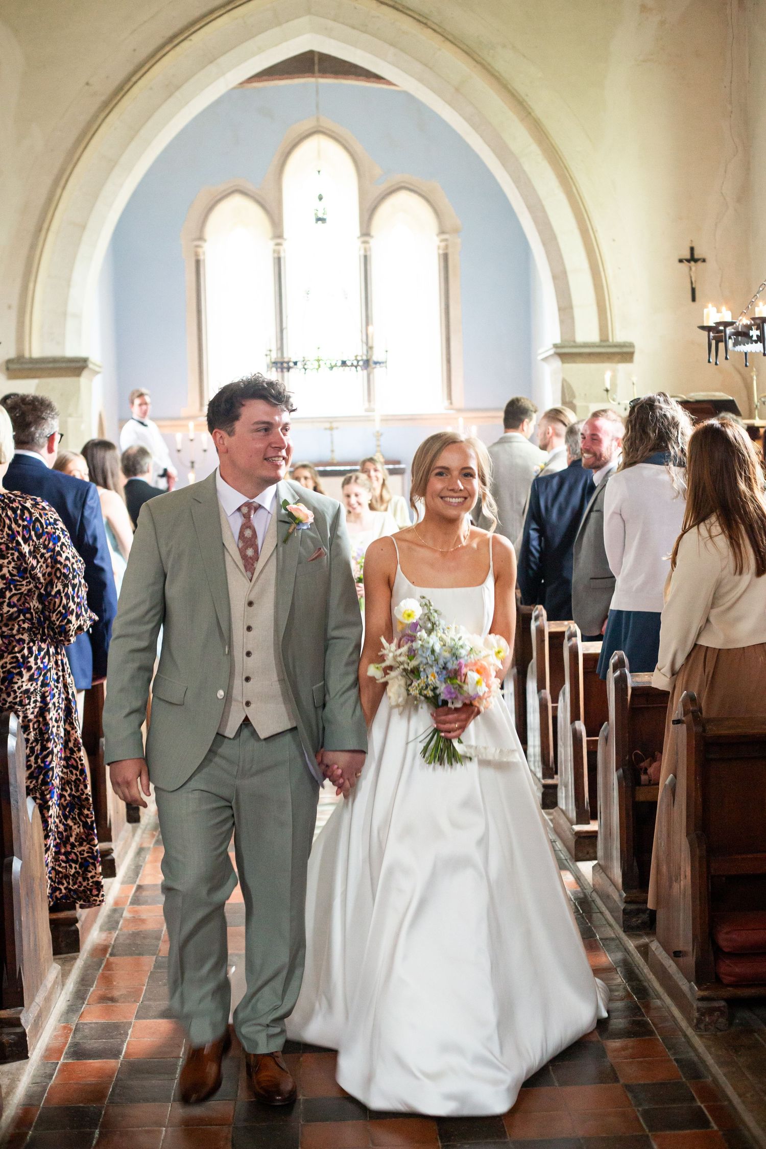 Newlyweds walk down the aisle of a beautiful church with arched windows and terracotta floors after their ceremony.