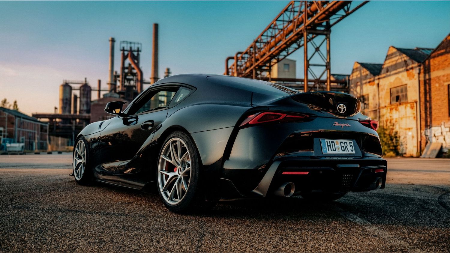 Sports cars photographed at sunset against industrial backdrop with orange crane structures.