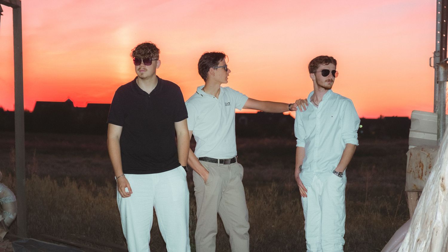 Three people stand together against a vibrant orange sunset sky in casual summer attire.
