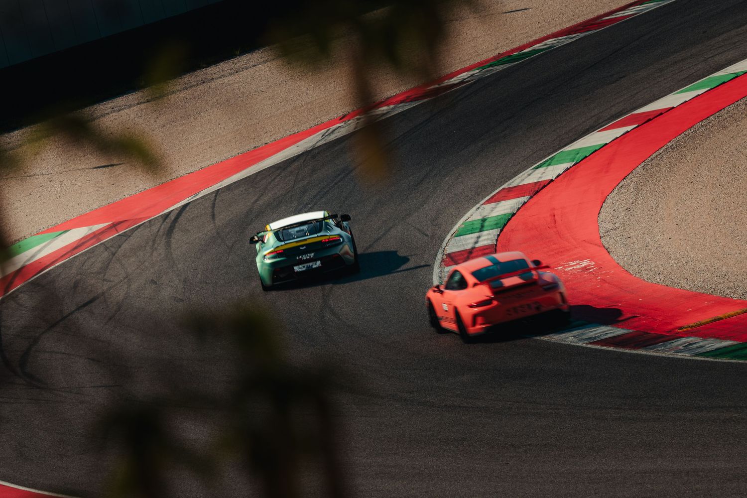 Two race cars, orange and green, competing on a red and white curbed racing circuit.