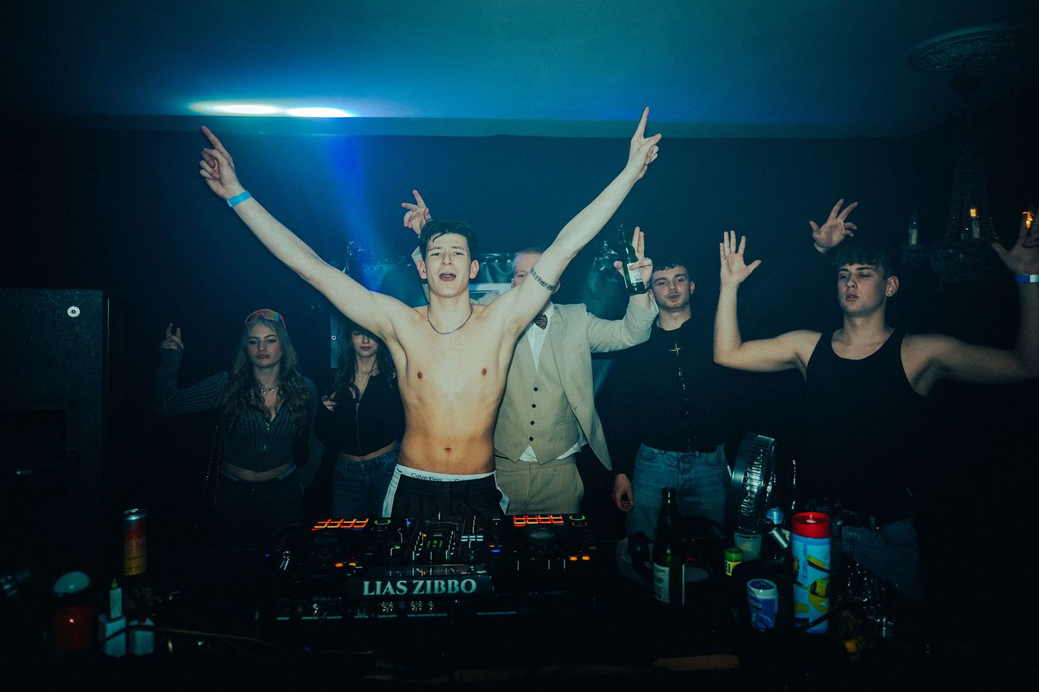 DJ performing at a nightclub with raised arms while energetic crowd dances under blue lighting and special effects.