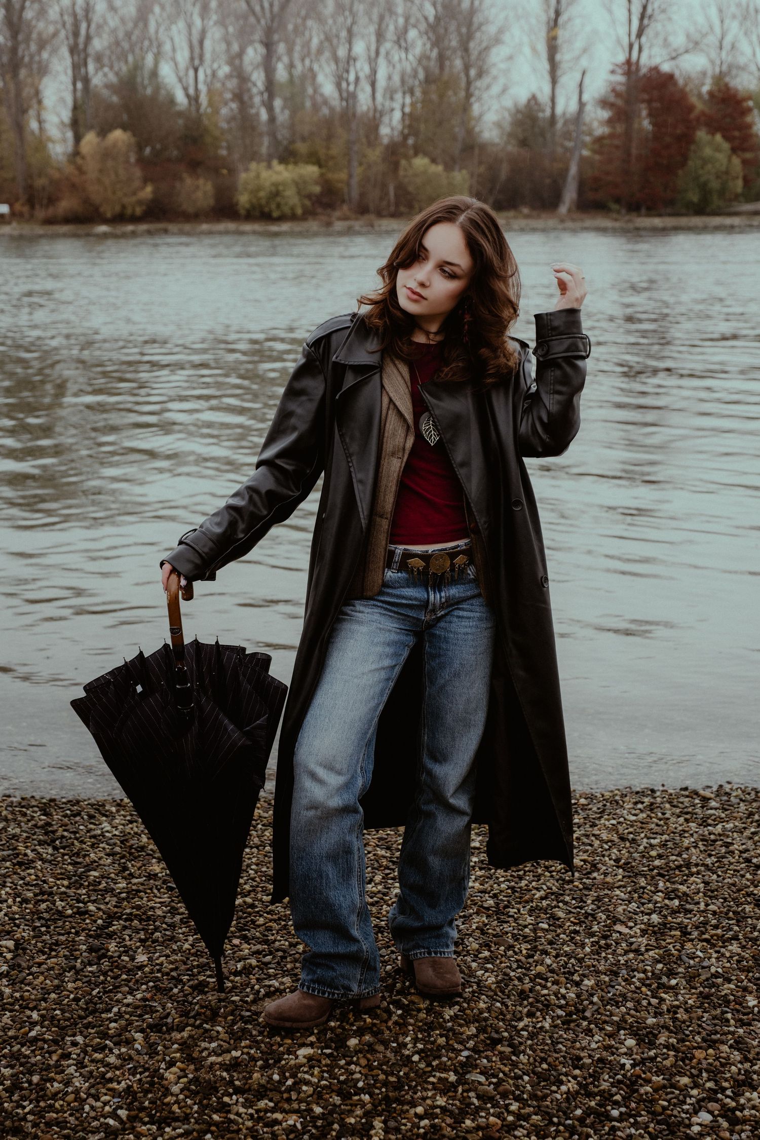 Figure with black umbrella posing on rocky riverbank in moody weather.