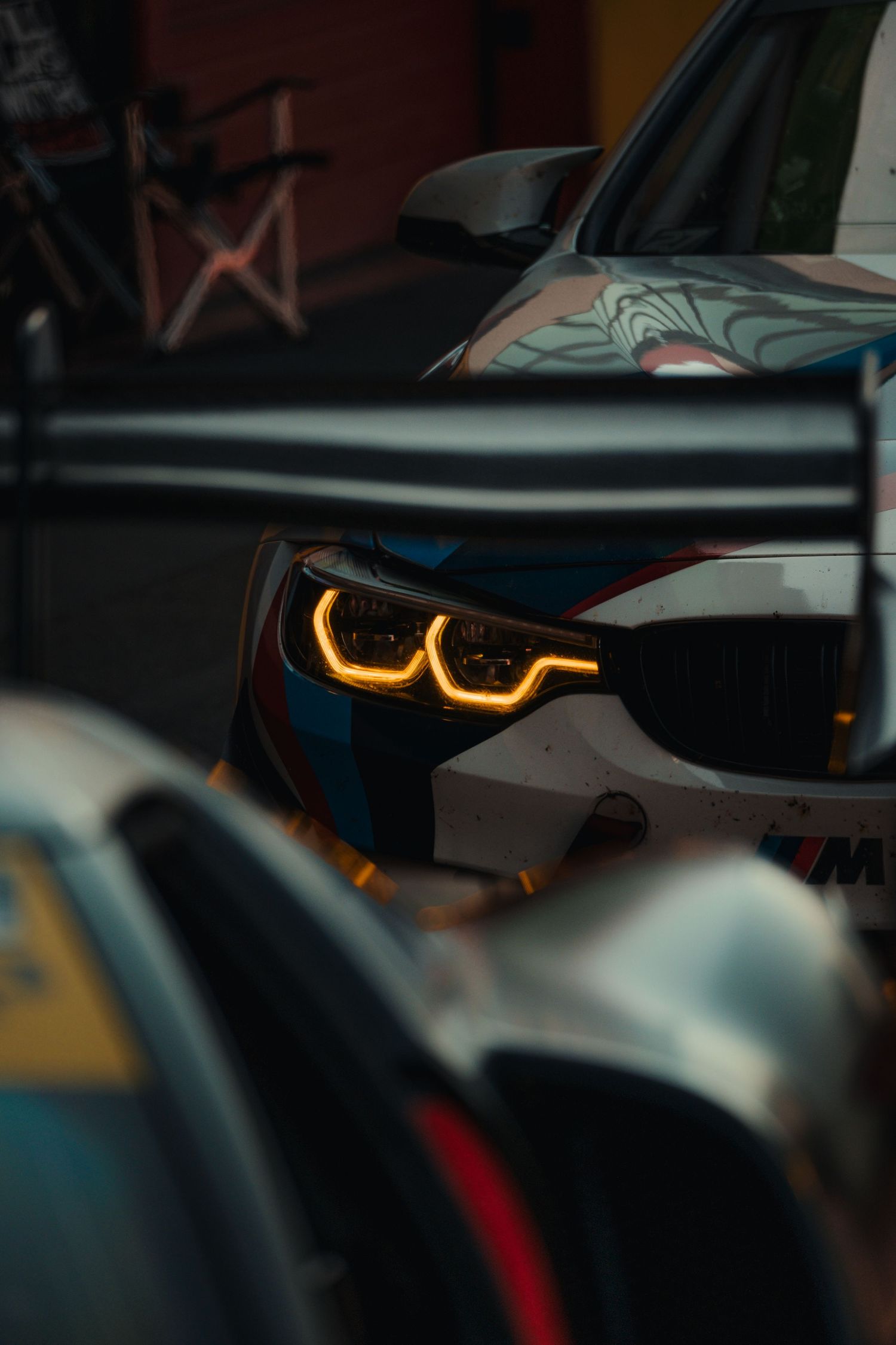 Close-up of a BMW M racing car headlight glowing amber in a pit lane, showcasing its distinctive kidney grille design.