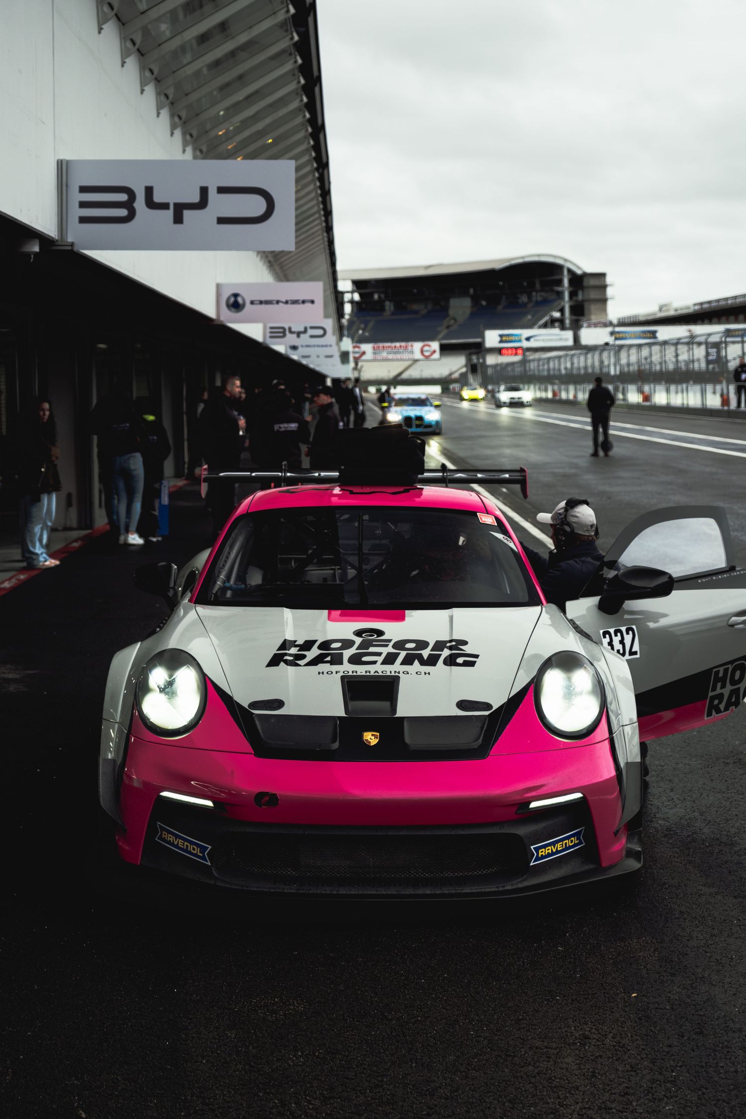 Pink and black Porsche race car with custom livery parked at a racing circuit under cloudy skies.