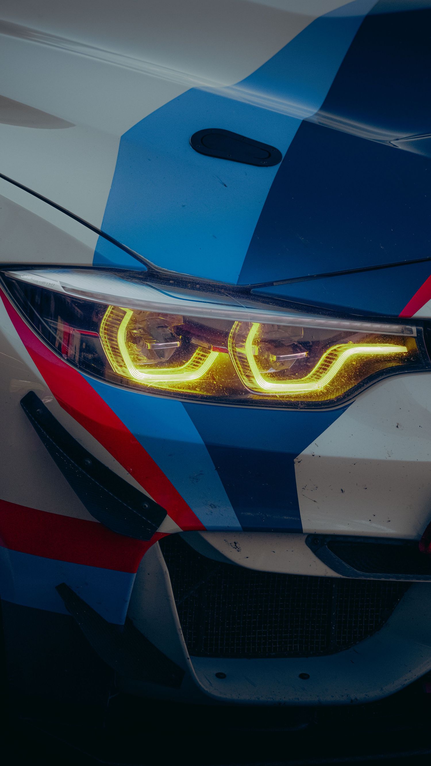 Close-up of a BMW M car headlight with glowing yellow LED DRL, featuring blue, white, and red M motorsport livery.