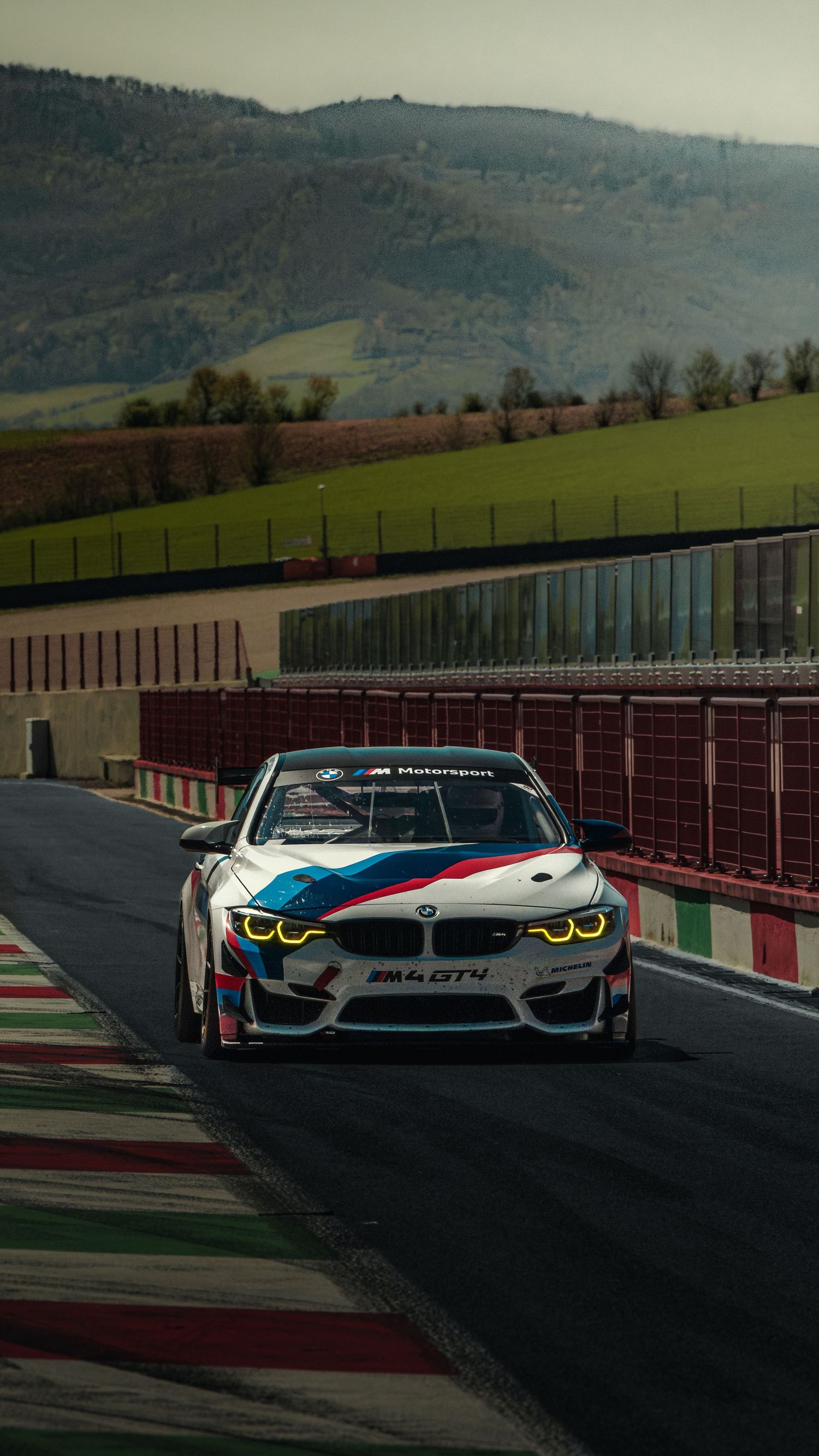 BMW M4 GT4 race car with M Motorsport livery driving on a circuit with green hills behind.