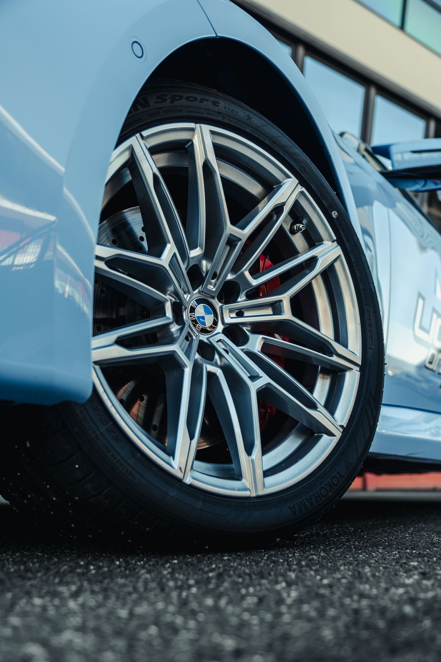 Close-up of a BMW alloy wheel with red brake calipers on a light blue BMW car parked on asphalt.