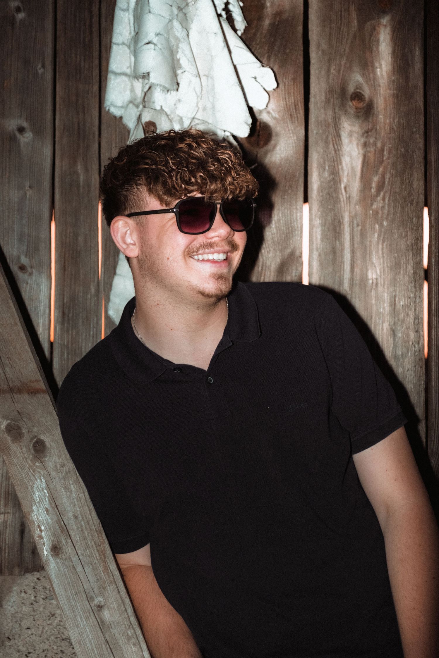Someone wearing sunglasses and a black polo shirt smiles while standing against a wooden wall in dim lighting.