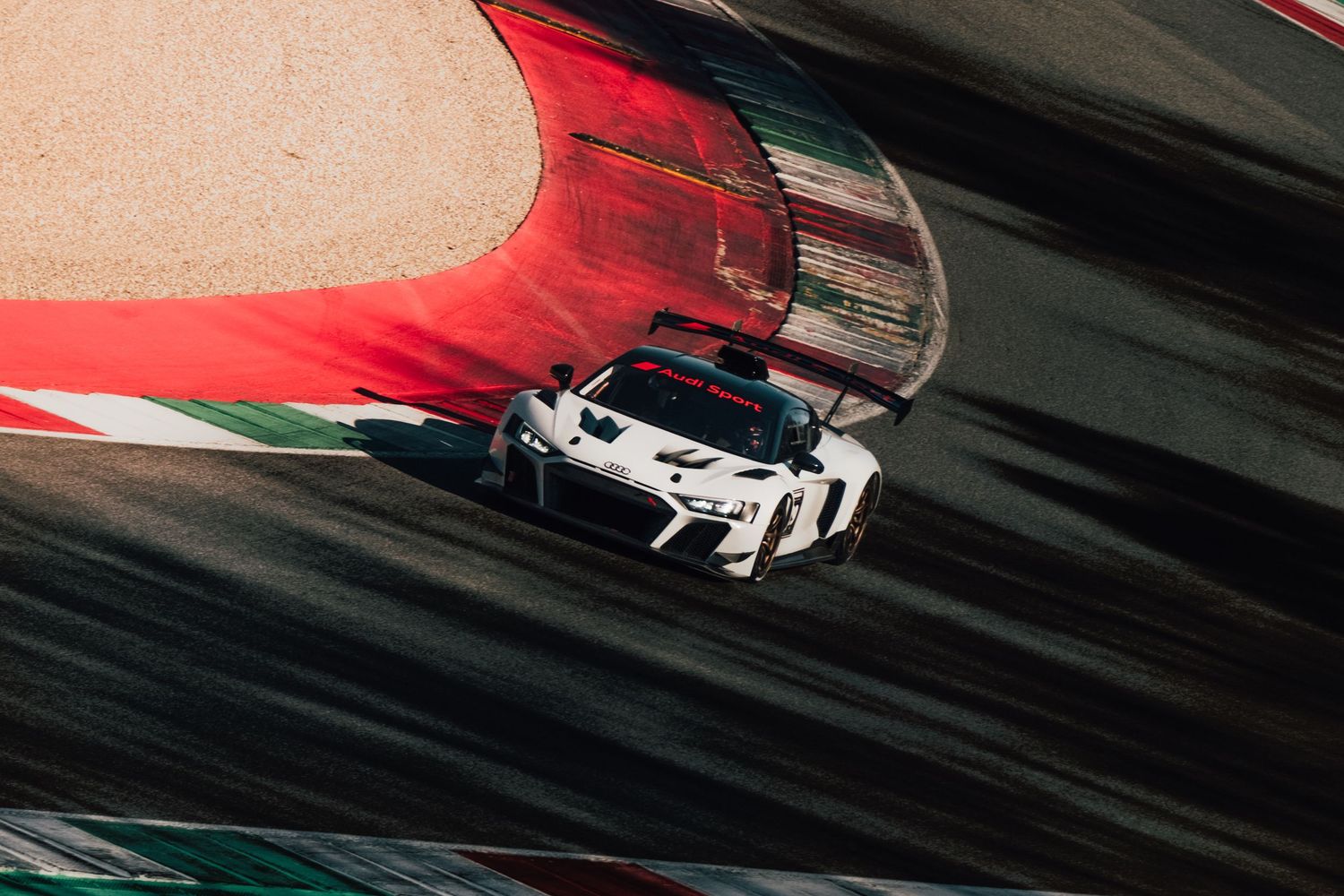 White Audi GT racing car speeding through a red and white barrier corner on a race track.