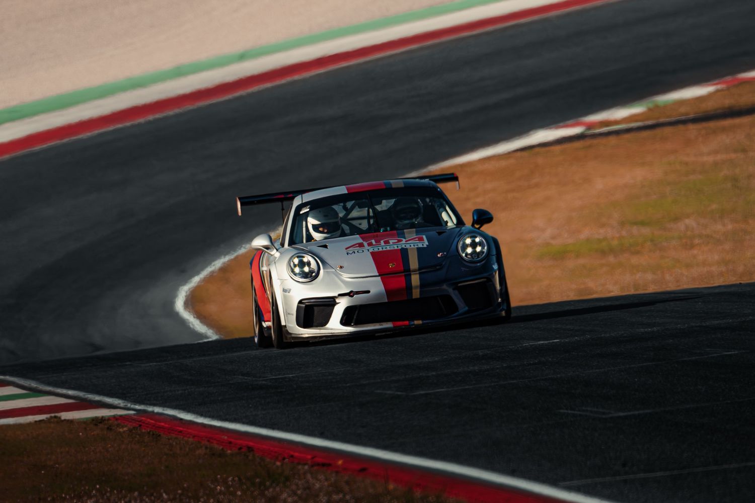 Porsche 911 GT3 Cup race car with red stripes navigating a corner on a racing circuit.