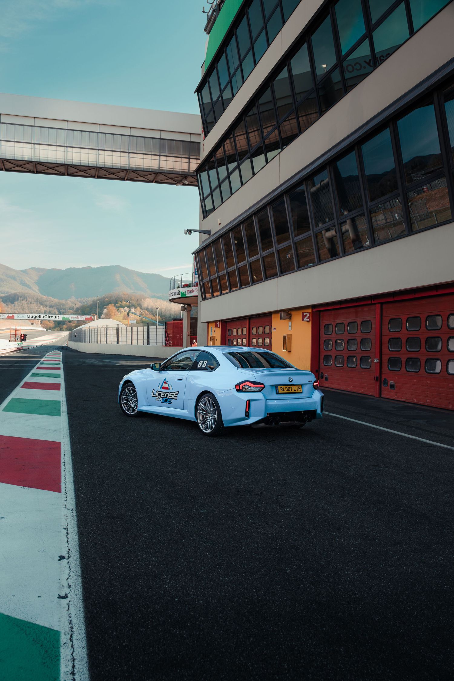Light blue BMW M2 sports car parked beside pit lane garages at a sunny racing circuit with mountain backdrop.