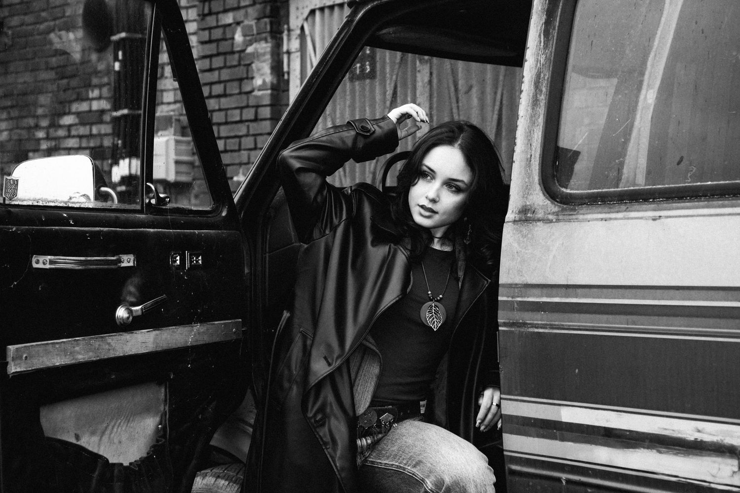 Dramatic black and white portrait against a brick wall with moody lighting and vintage car in background.