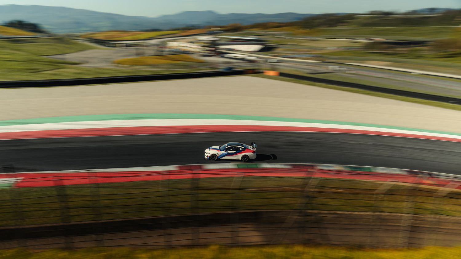 Aerial view of a white race car speeding along a scenic motorsport circuit track.
