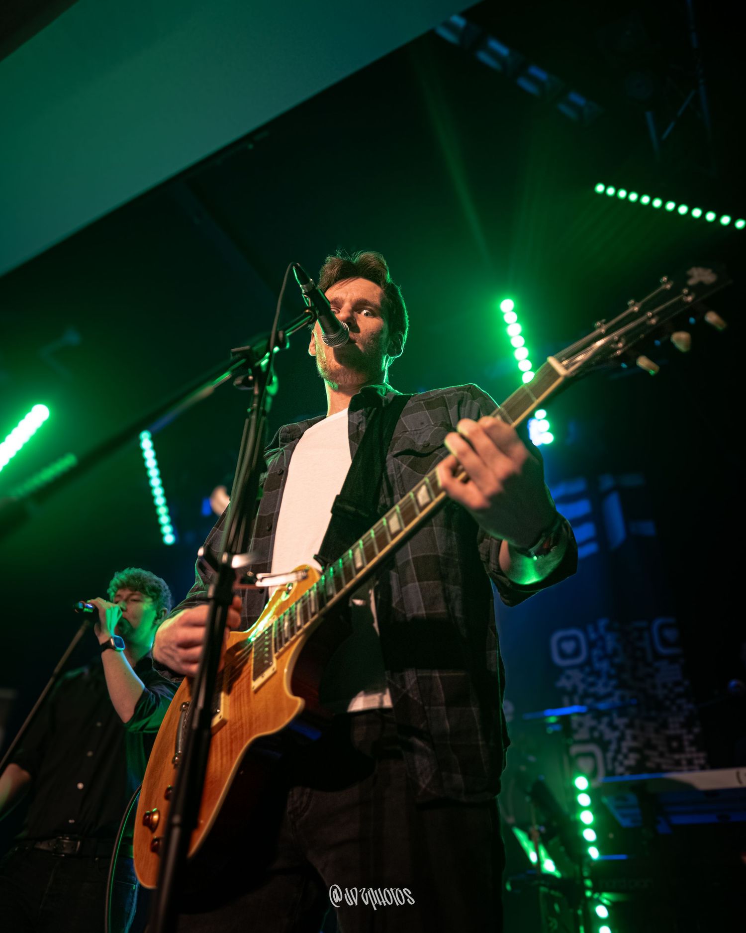 Concert performance with guitar players silhouetted against green and blue stage lighting.