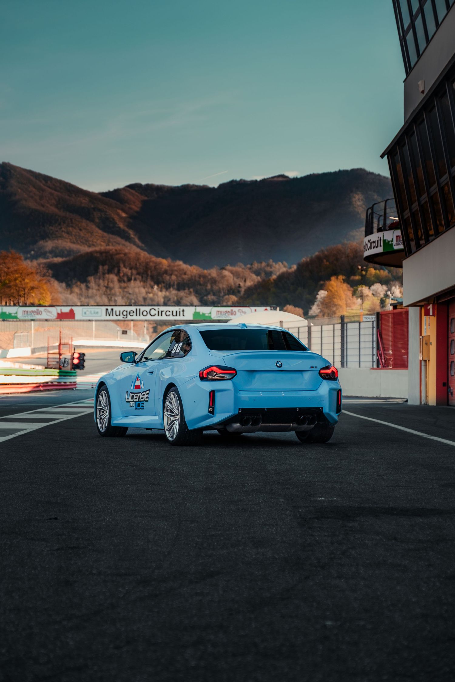 Light blue BMW M2 race car at Mugello Circuit with mountain backdrop.