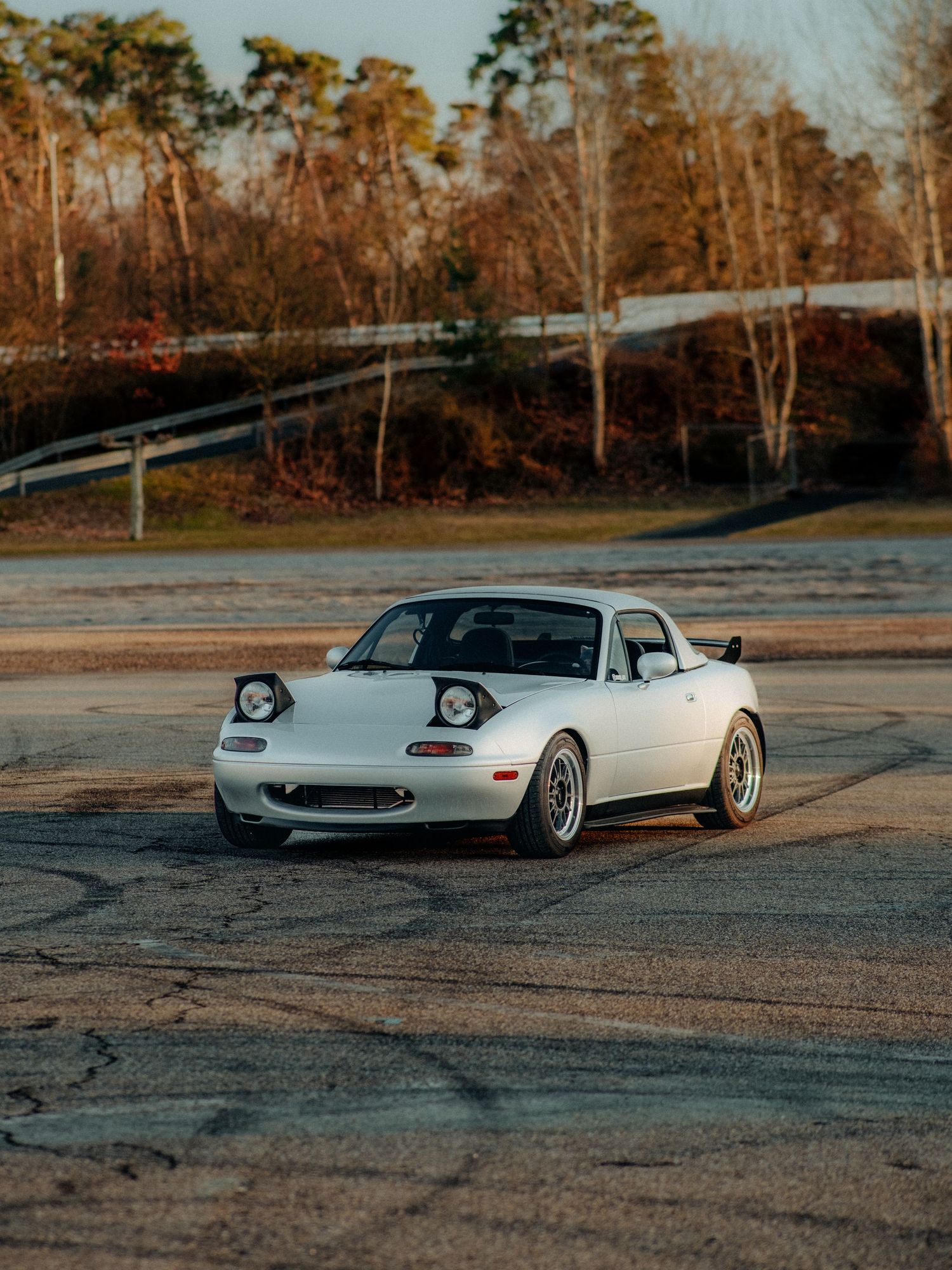 White Mazda Miata with custom wheels parked in a lot at sunset with autumn trees behind.