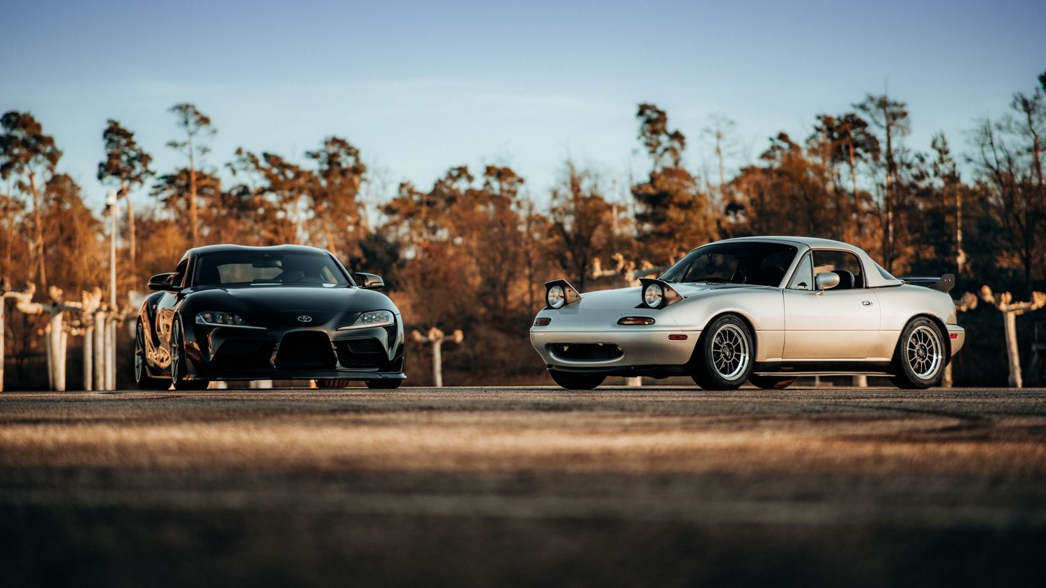 Black Toyota Supra and white Mazda Miata parked together at sunset with trees in background.
