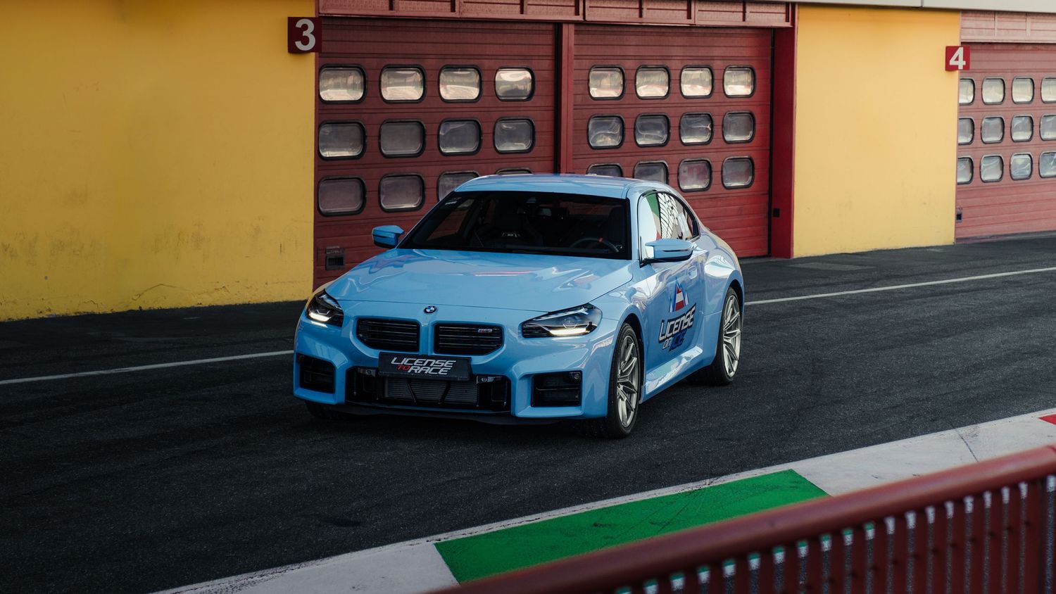 Light blue BMW M2 race car parked in front of red garage doors on a racing circuit.