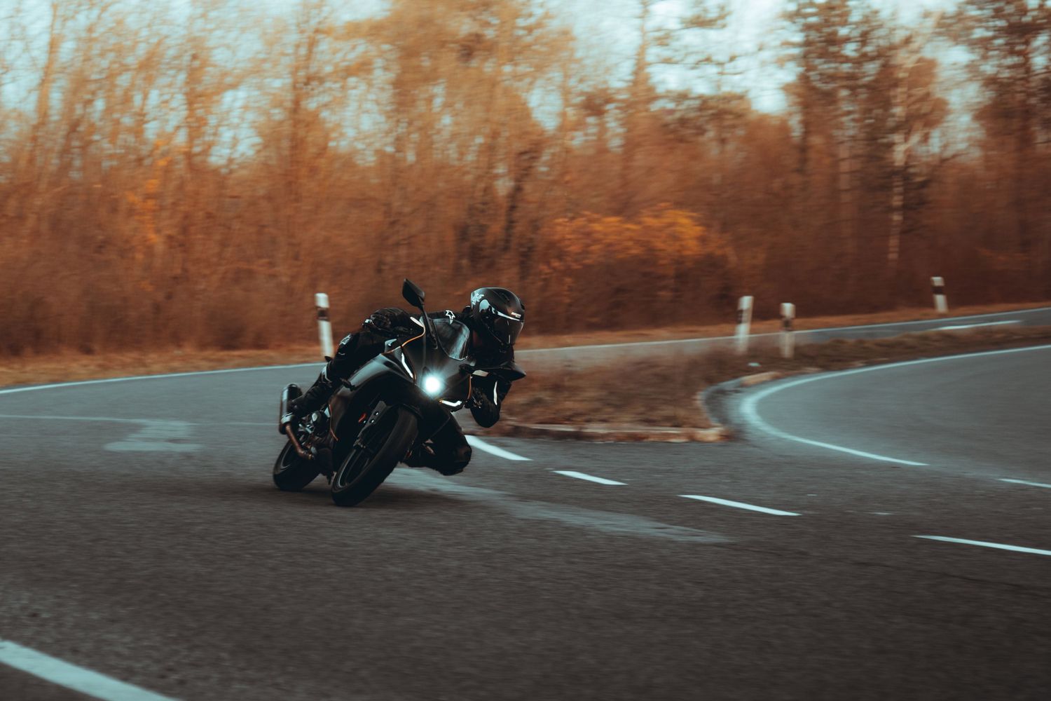 A motorcycle rider leans into a curve on a winding mountain road during sunset with autumn trees in the background.