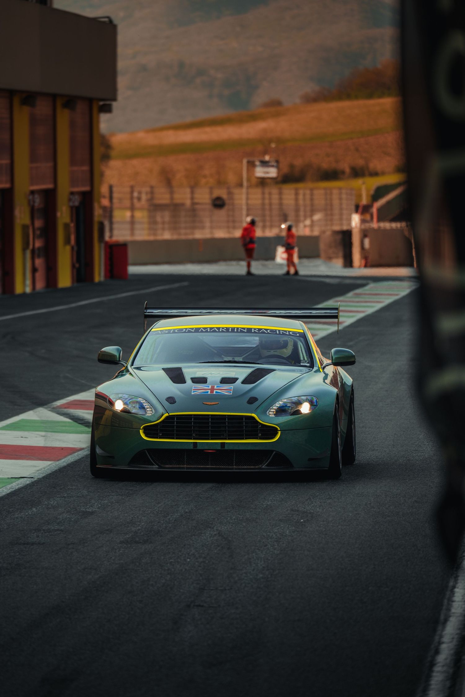 Green Aston Martin Vantage GT4 race car exiting pit lane on a motorsport circuit.