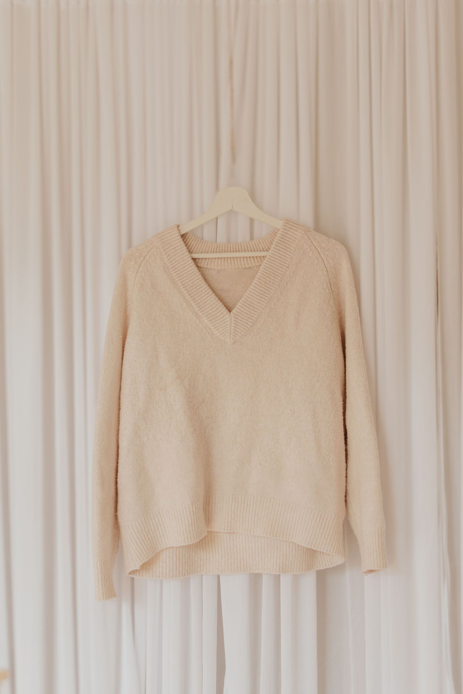 Ein gemütlicher cremefarbener Pullover mit V-Ausschnitt hängt an einem weißen Holzbügel vor einem hellen Vorhanghintergrund.