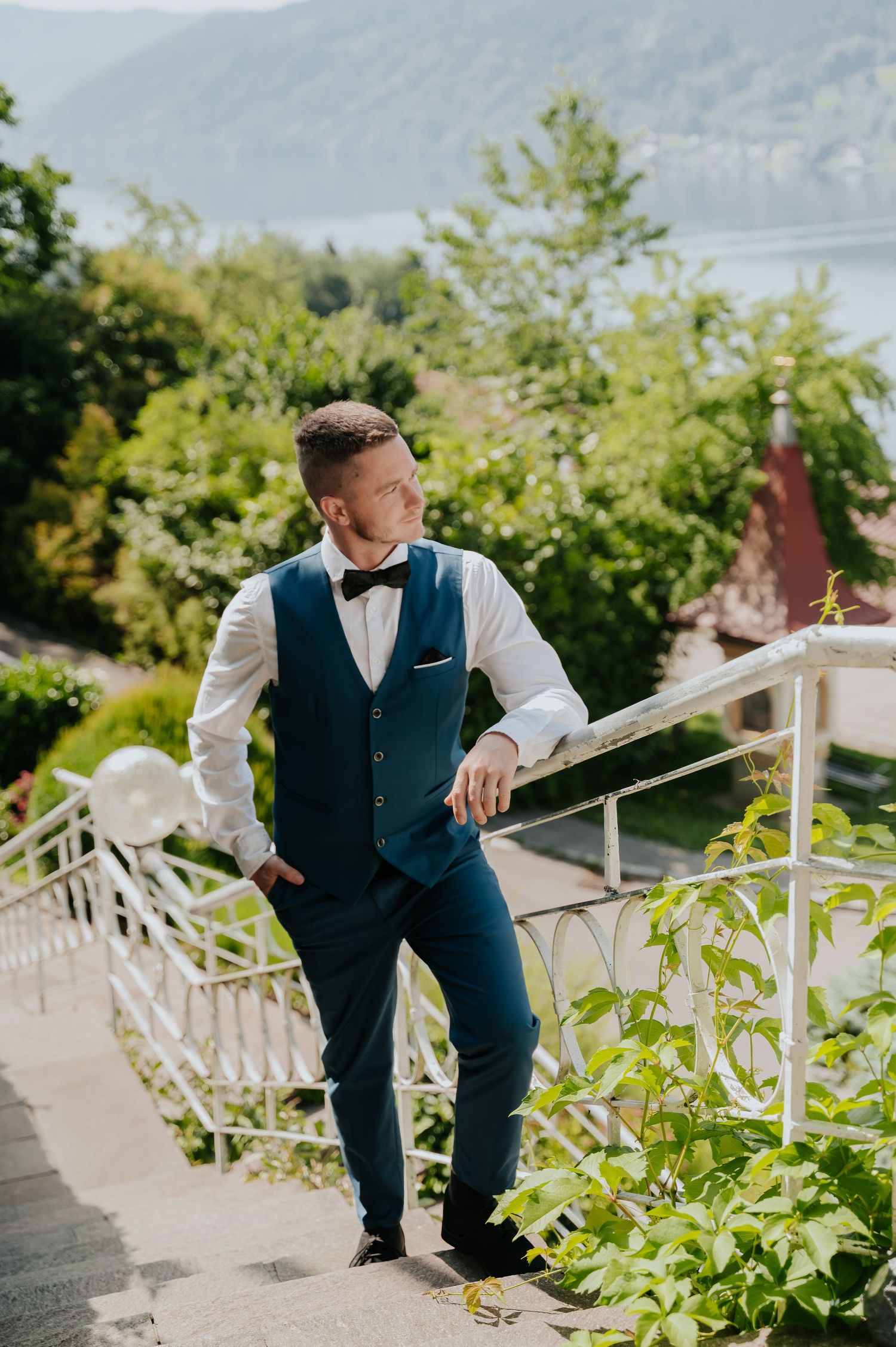Elegant groom in blue vest and bow tie posing on outdoor stairs with lush greenery and lake view.