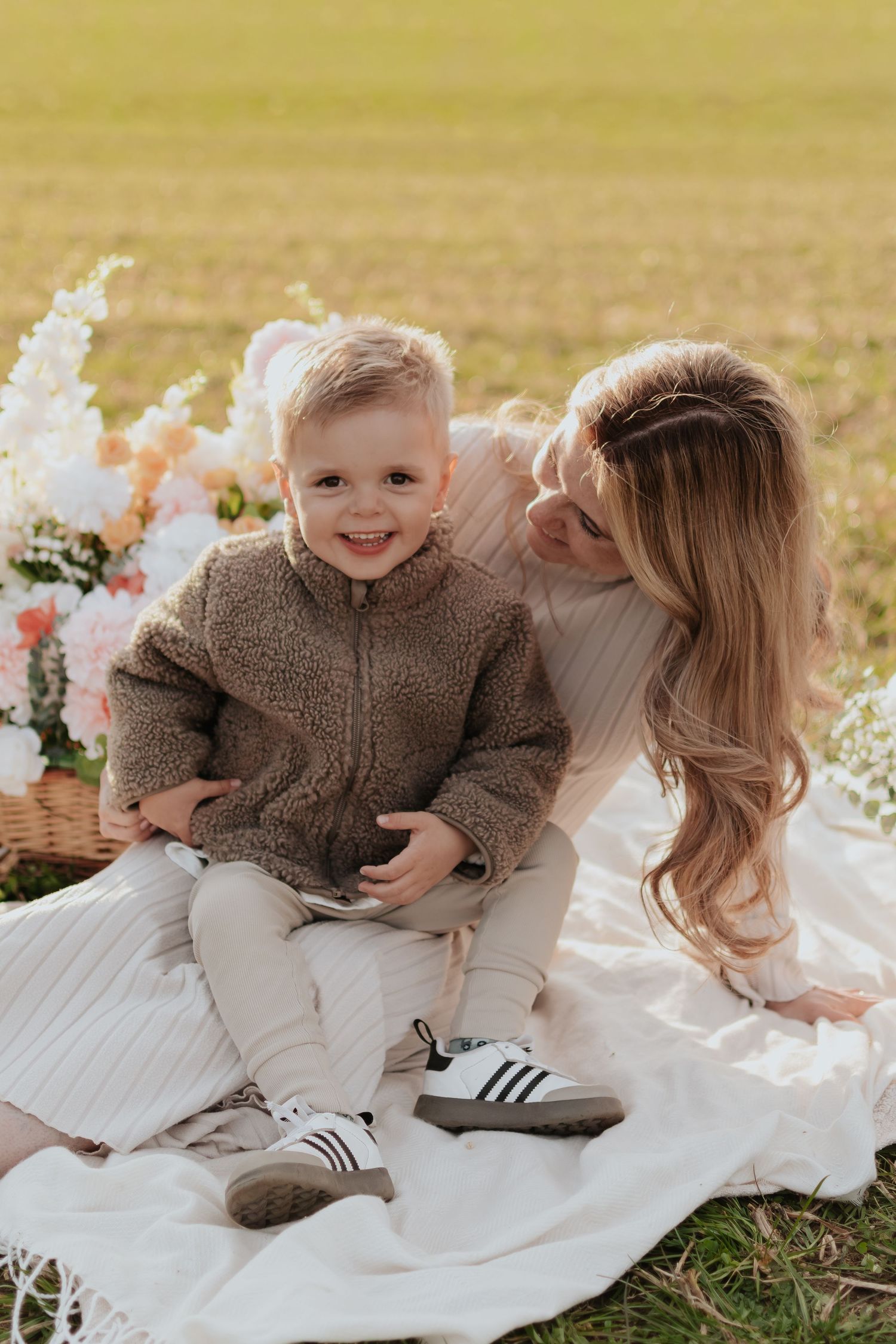 A smiling toddler boy in a brown fleece jacket sits on a blanket outdoors with his mother during a sunny picnic.