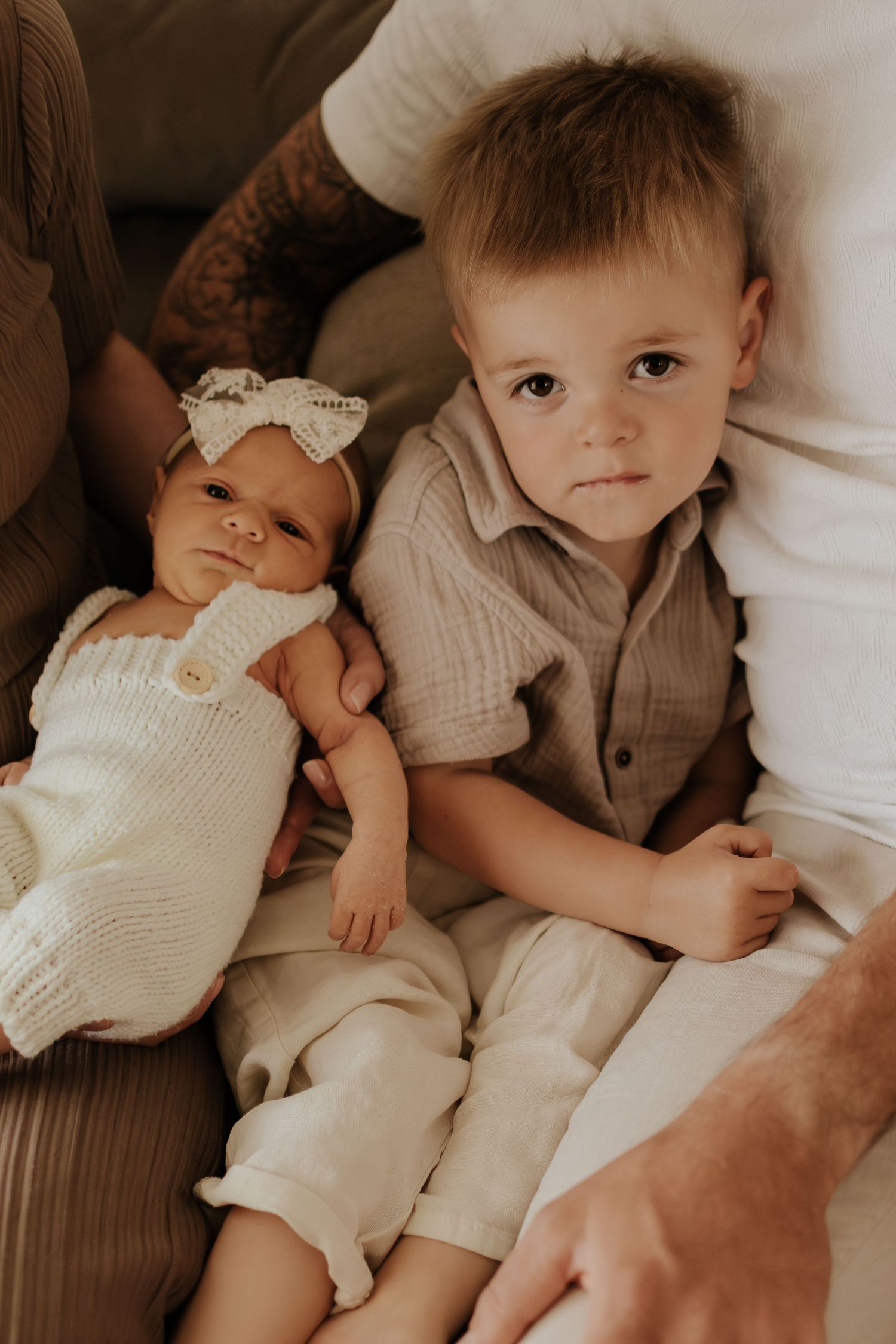 A toddler boy in beige linen sits beside a newborn baby girl in a white knit romper with a lace bow headband.