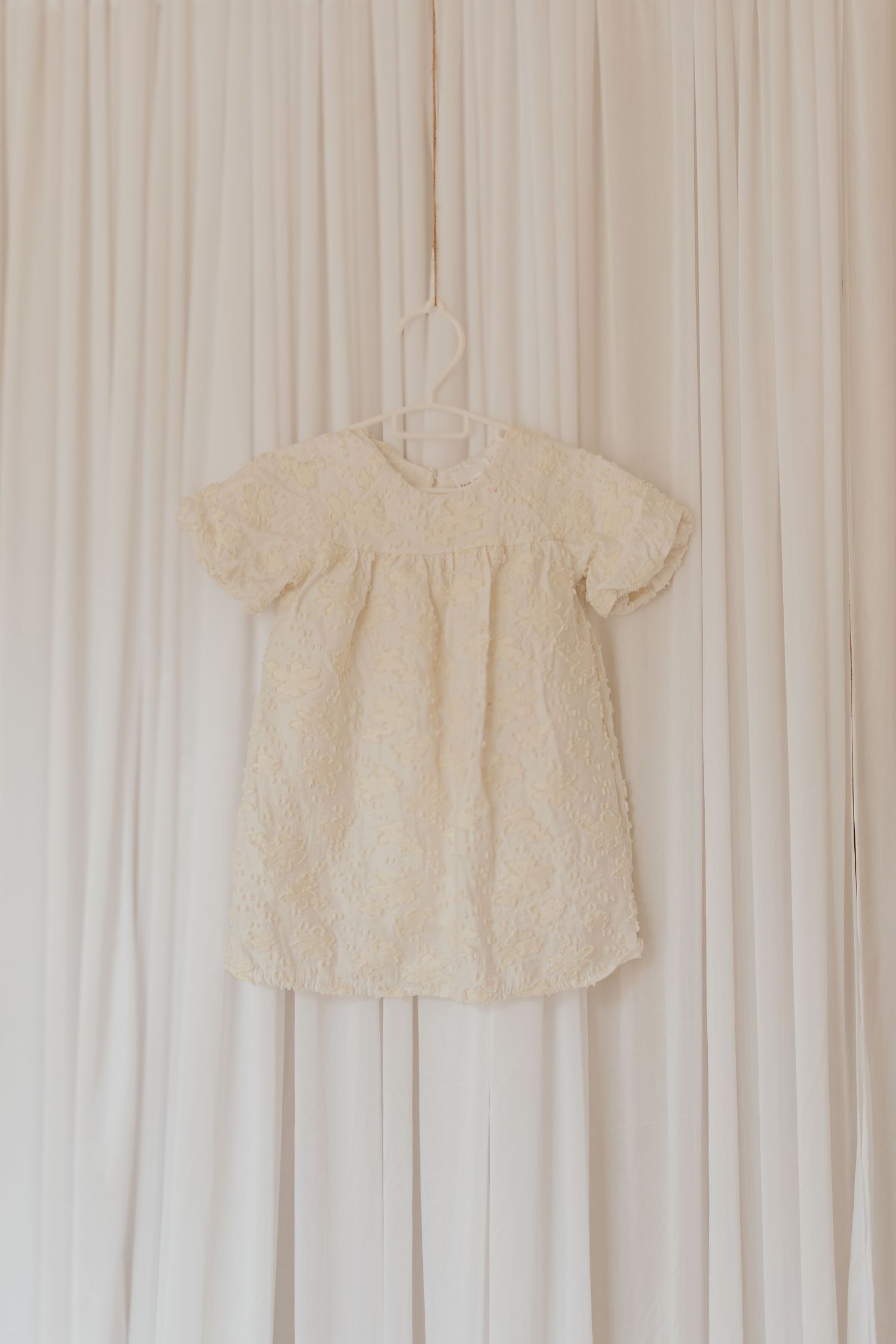 Vintage weiße Baumwoll-Babykleider, die an einer Wand vor einem hellen cremefarbenen Hintergrund hängen.