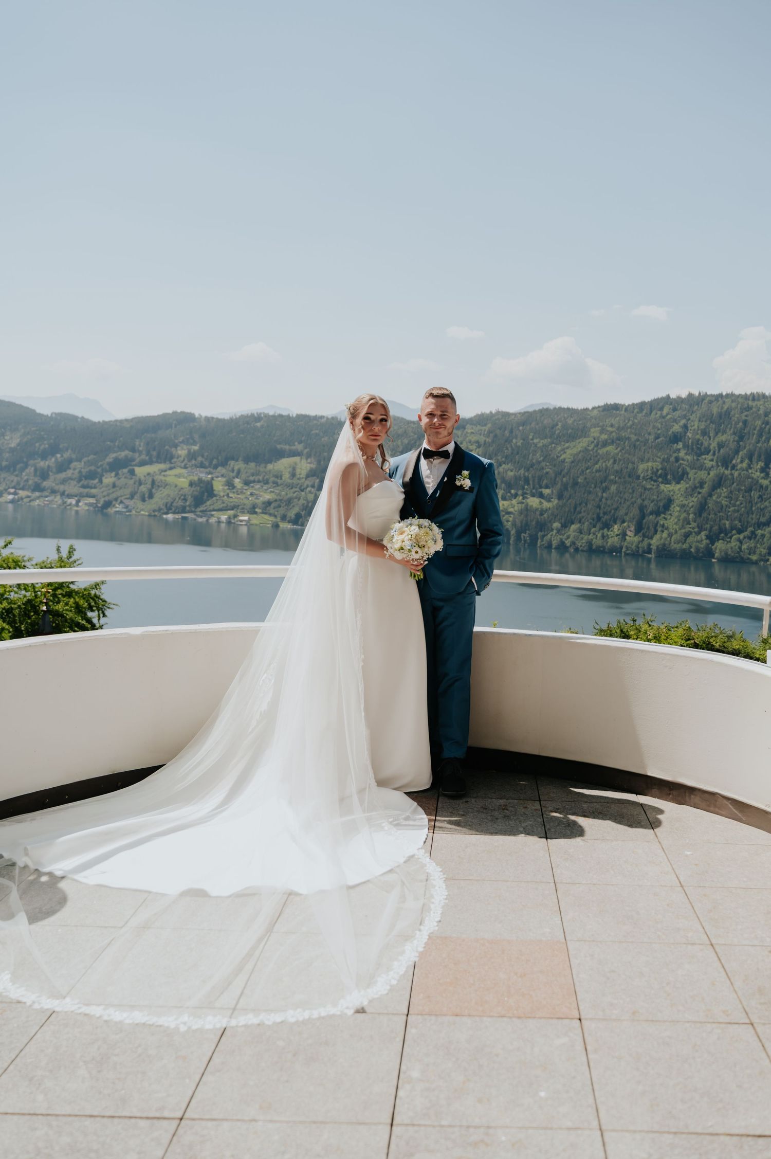 Bride and groom posing on a scenic terrace overlooking a lake and green hills on their wedding day.