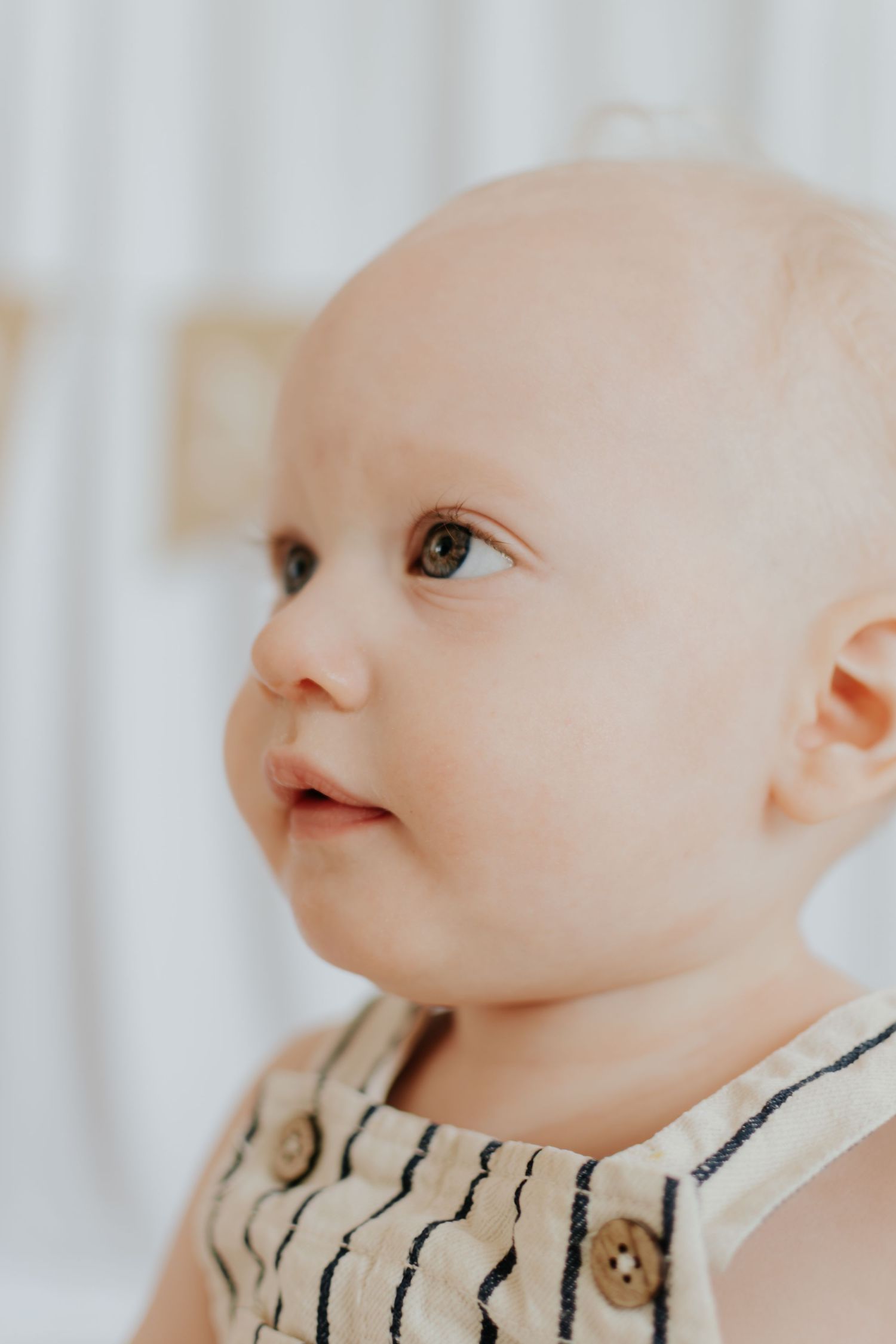 Adorable baby in a striped linen romper with wooden buttons, looking up curiously against a soft white background.