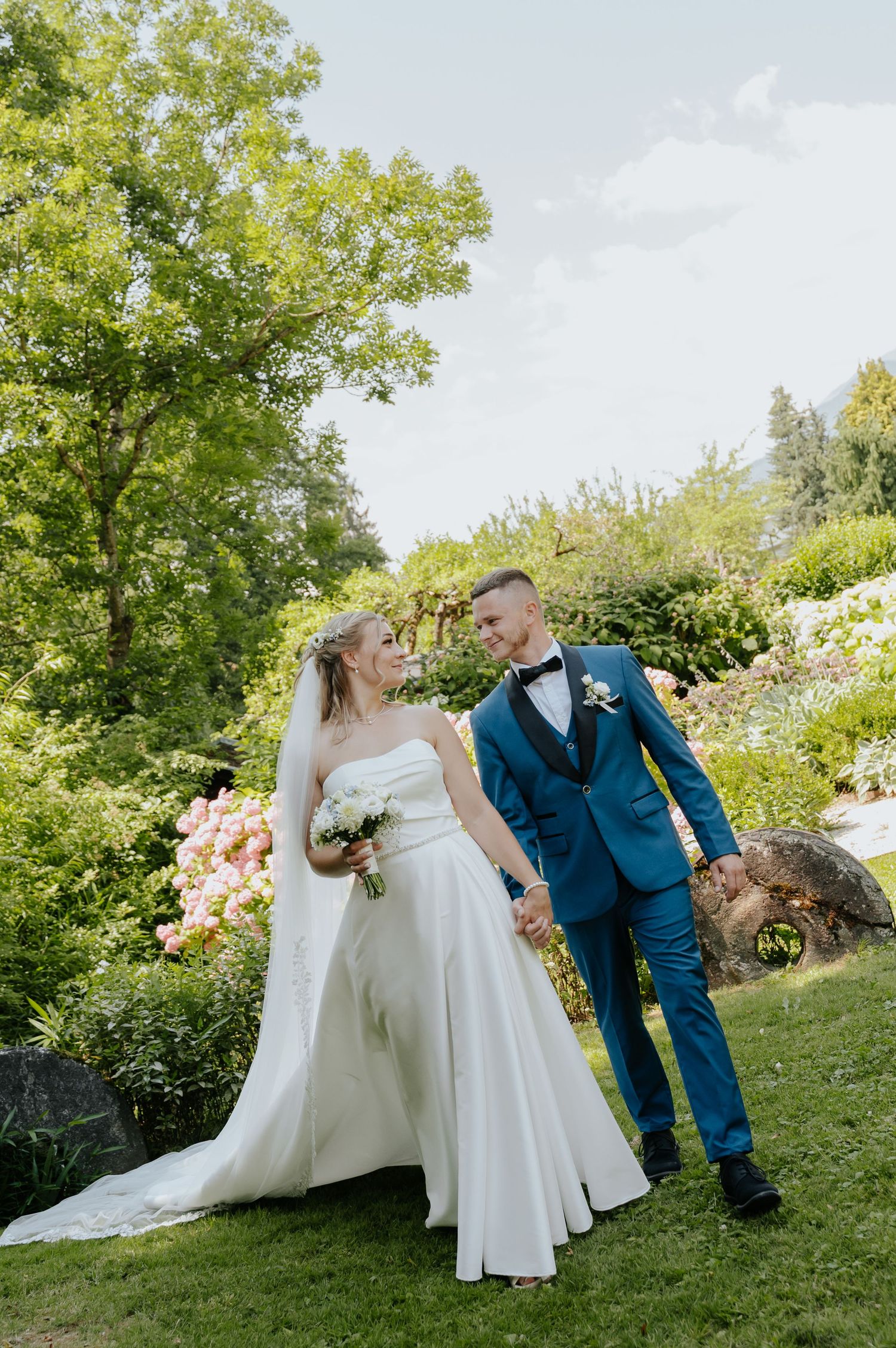 Bride in white gown and groom in blue tuxedo hold hands walking through lush garden on their wedding day.