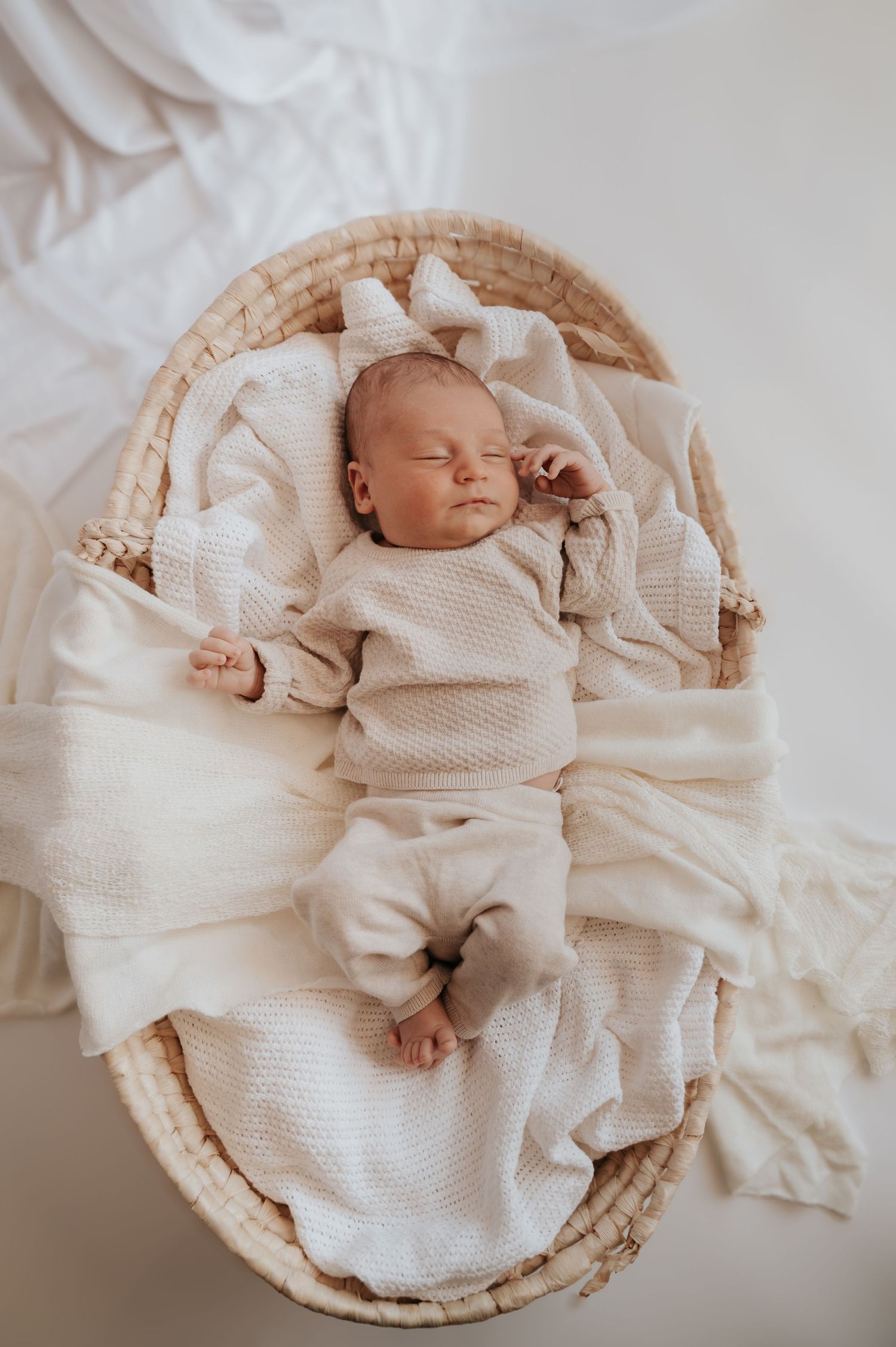 Newborn baby sleeping peacefully in a wicker moses basket, wrapped in cream knit blanket with a white bow headband.