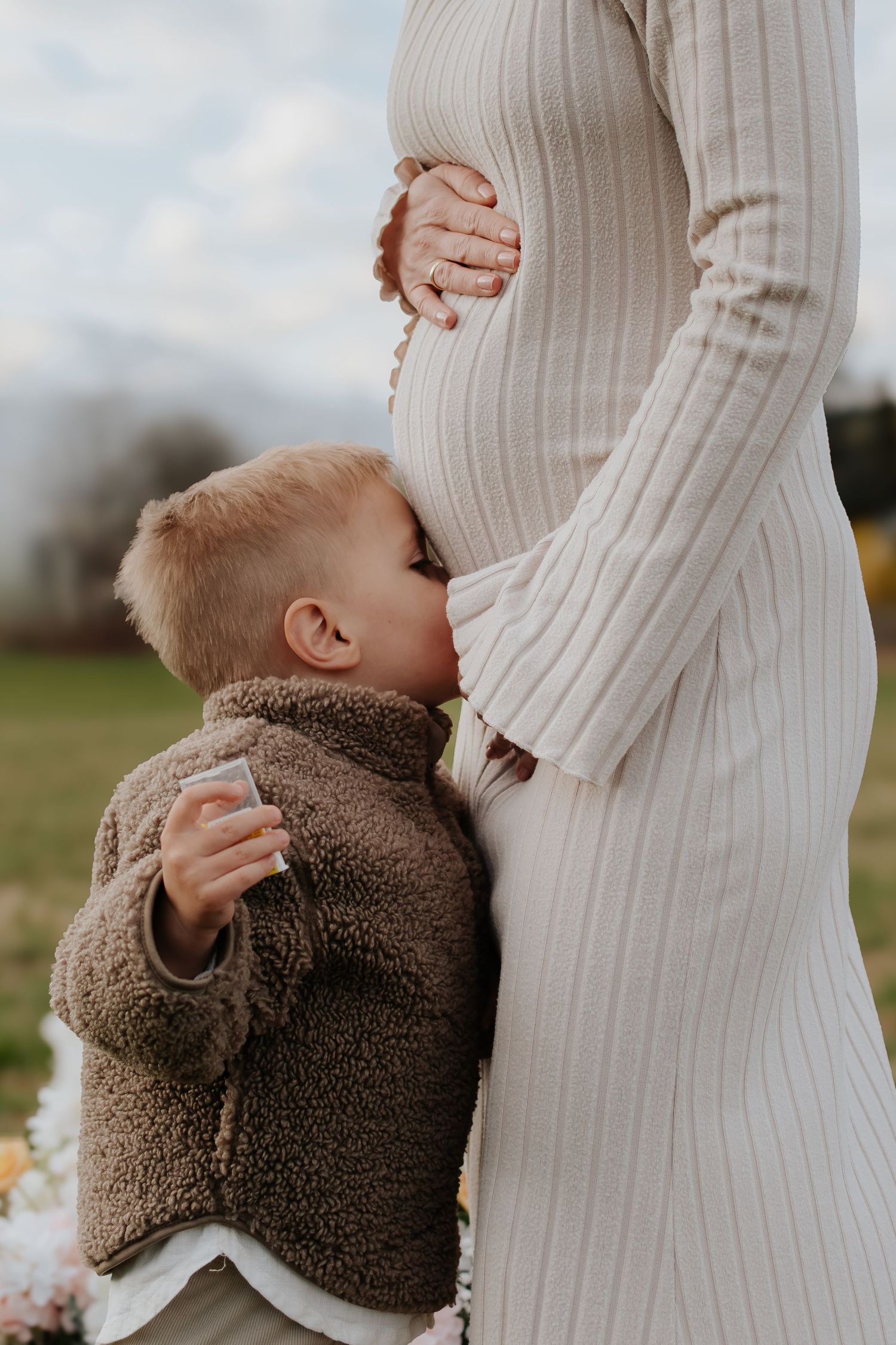 Young boy hugging pregnant mother's belly outdoors during a maternity photo session in a green field.