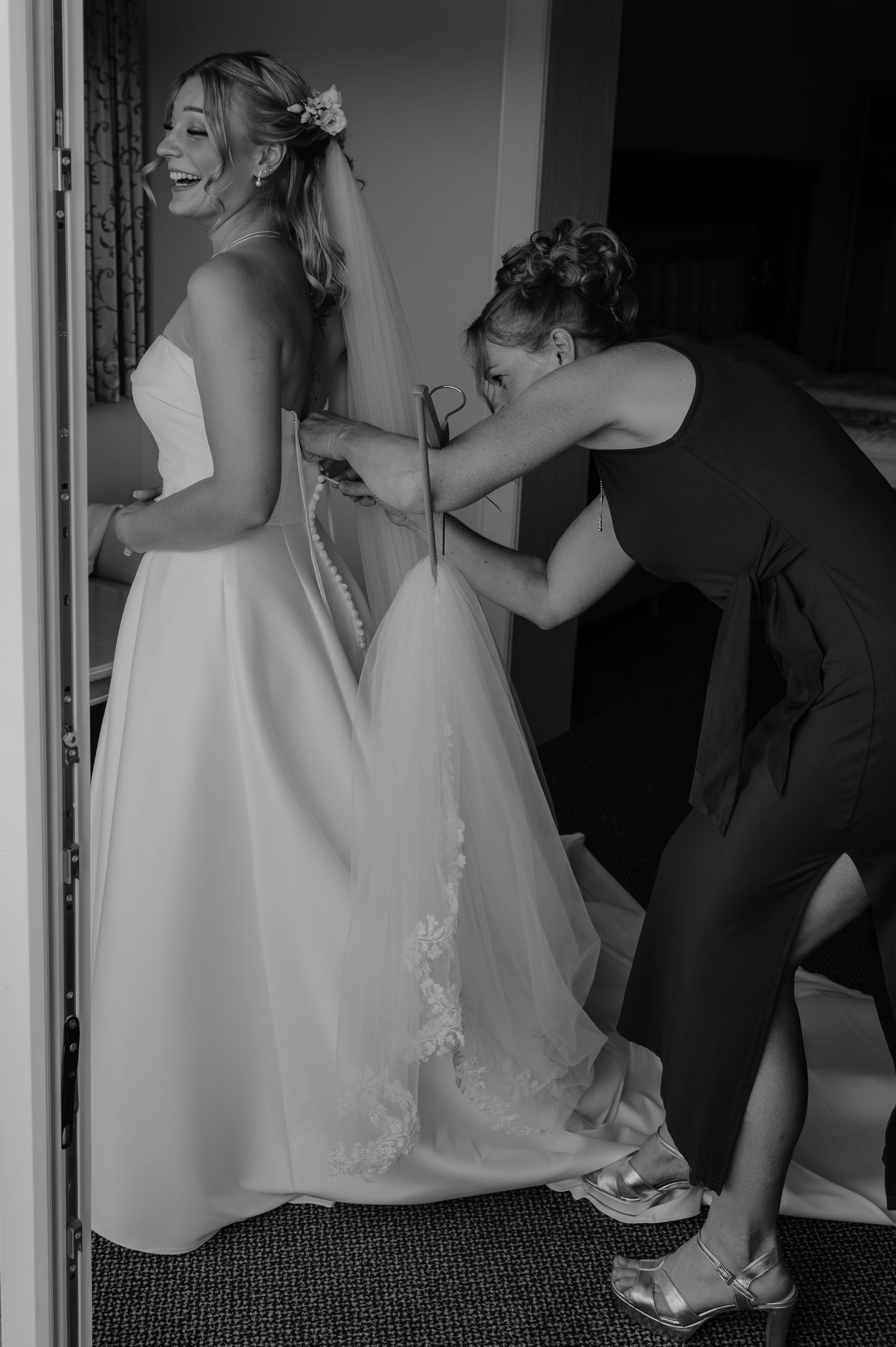Bridesmaid helping bride button up her elegant white wedding gown in a black and white photo.