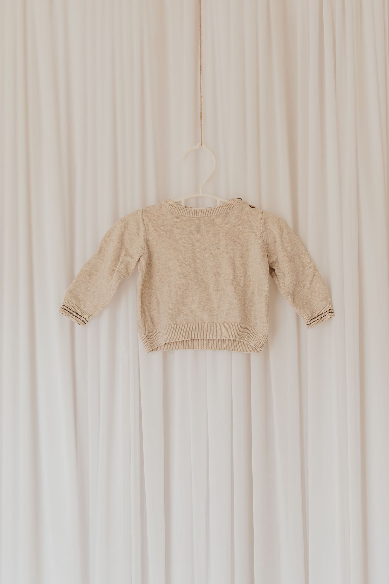 Ein beiger gestrickter Babypullover, der vor einem weißen Vorhanghintergrund in minimalistischem Styling hängt.