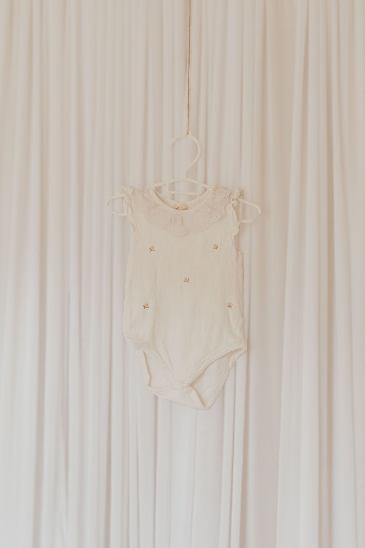 Weiße Baby-Strampler und Kleidungsstücke, die an einer weißen Gardinenstange vor einem hellen Hintergrund hängen.