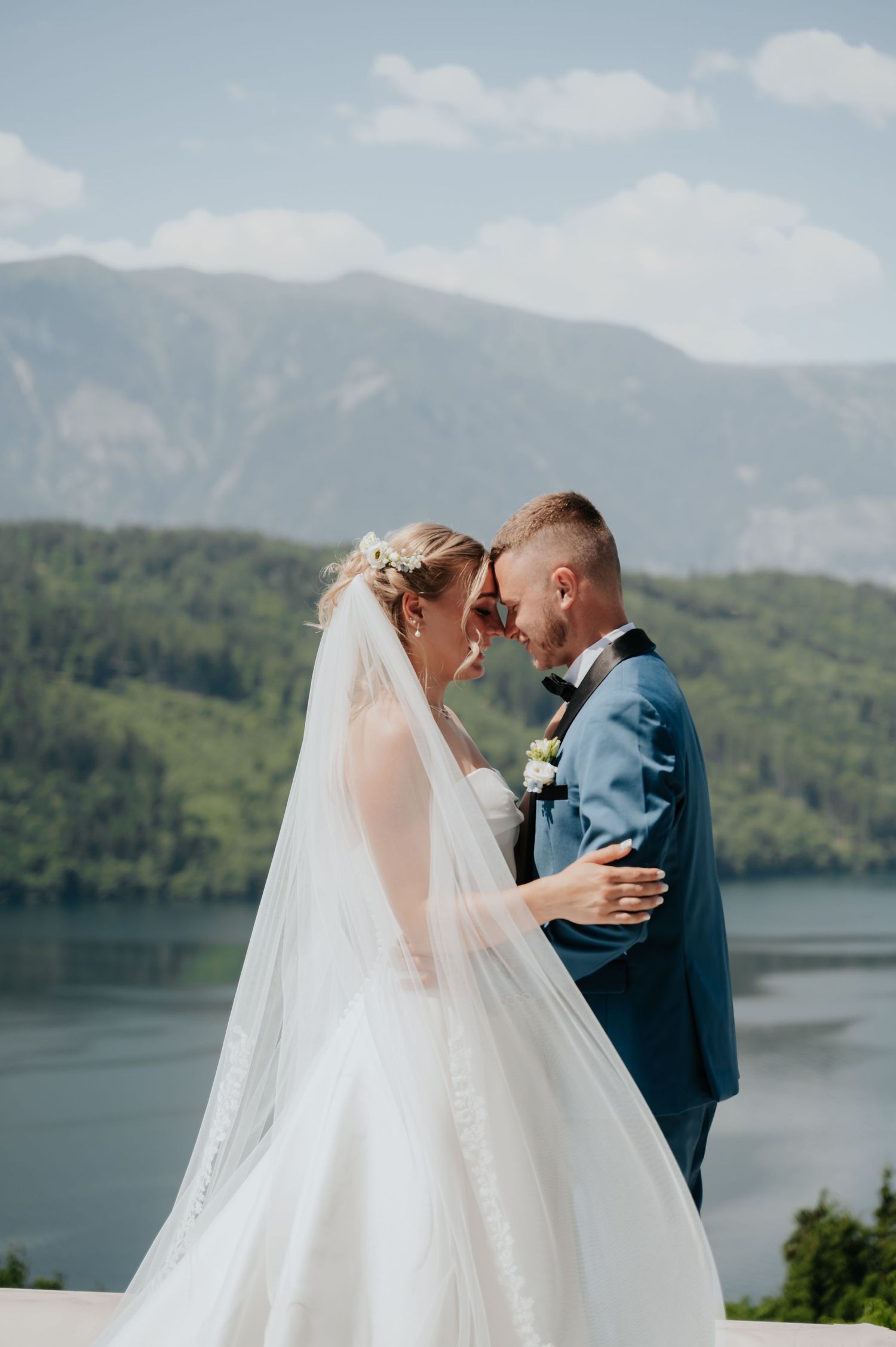 Bride and groom share romantic forehead touch in wedding attire with mountain lake backdrop on sunny day.