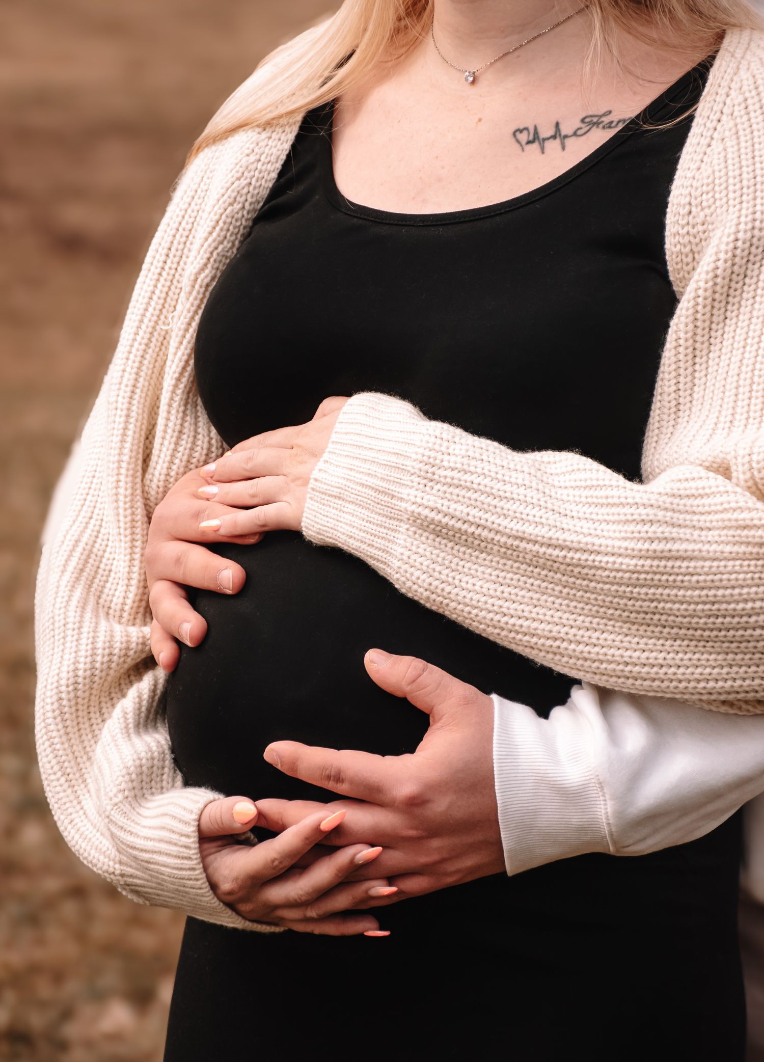 Nahaufnahmen von den Händen eines erwartenden Paares, die den Babybauch in einem cremefarbenen Pullover wiegen.