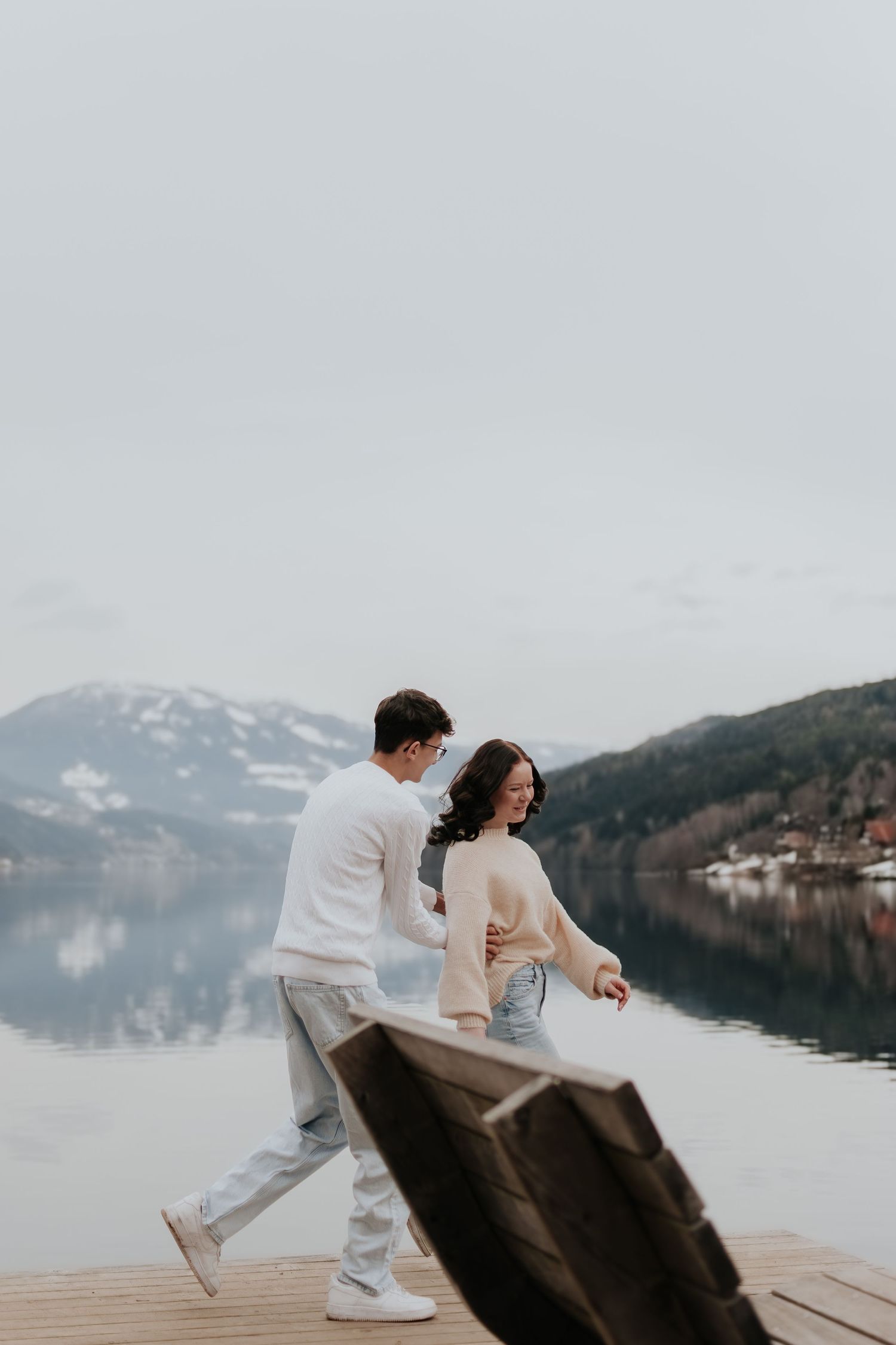 Couple walking on a wooden dock by a serene alpine lake with snow-capped mountains in the background.
