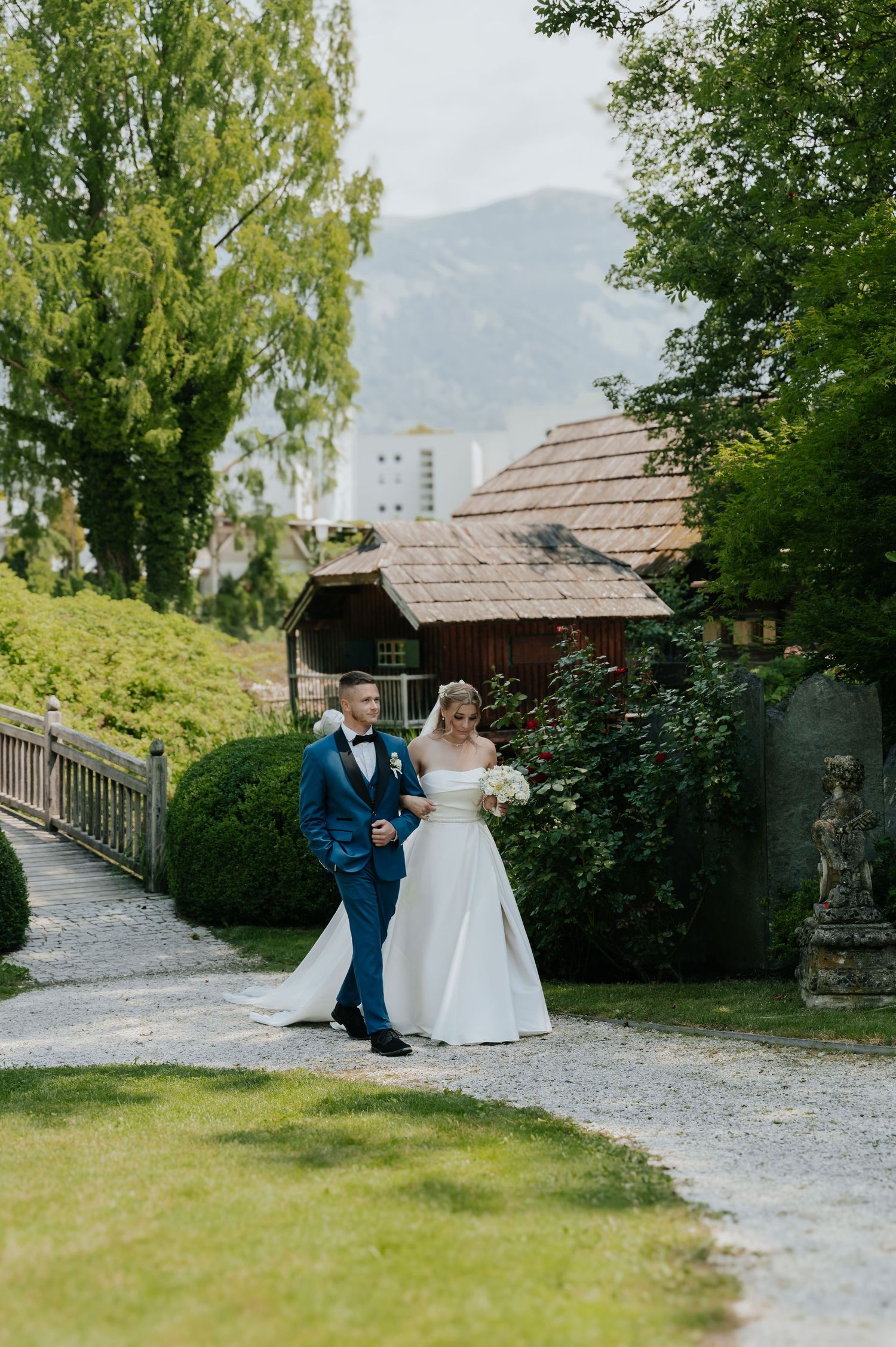 Bride and groom walk hand-in-hand along a garden path with mountains and a charming cottage in the background.