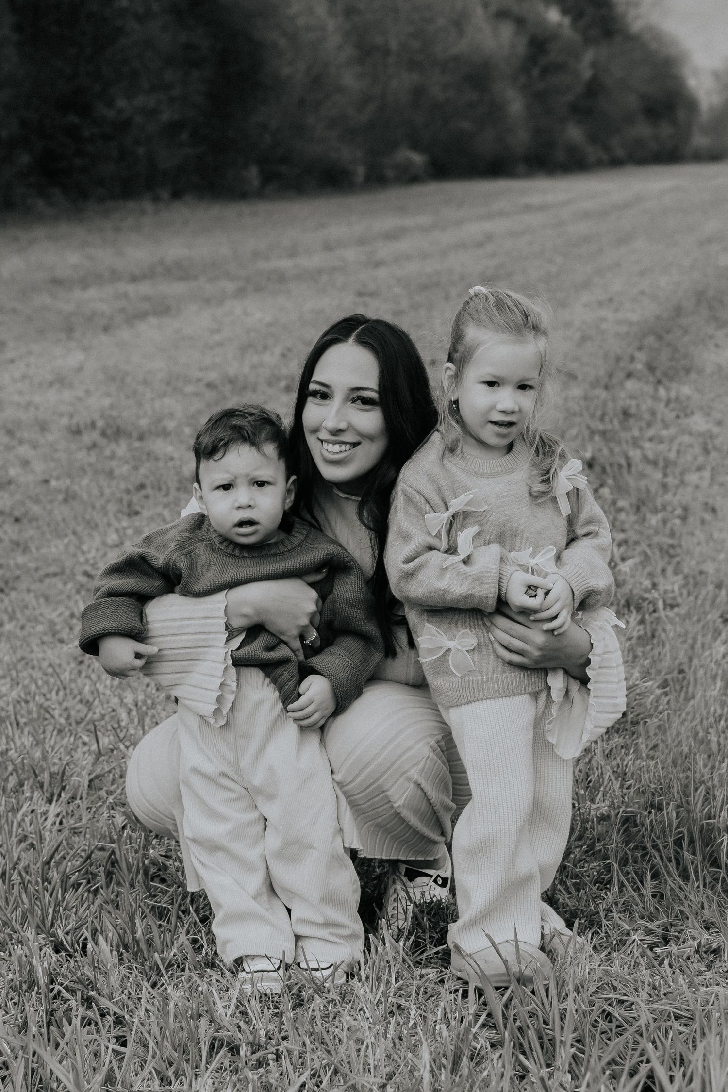 Black and white photo of a smiling mother crouching in a field with her two young children.