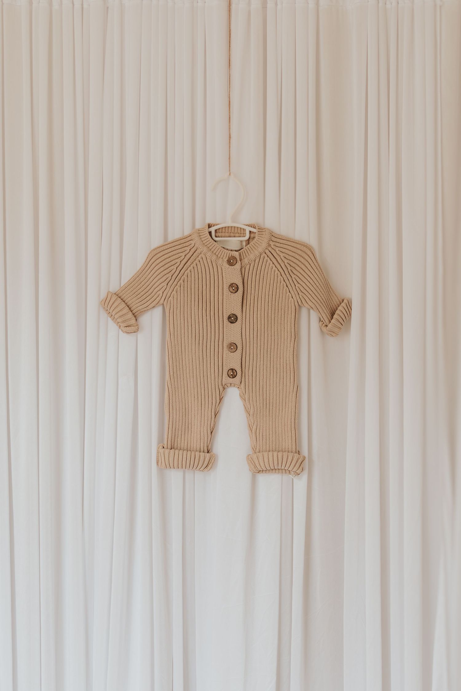 Beige gerippte Strickjacken für Babys, die an einer weißen Holzwand hängen.