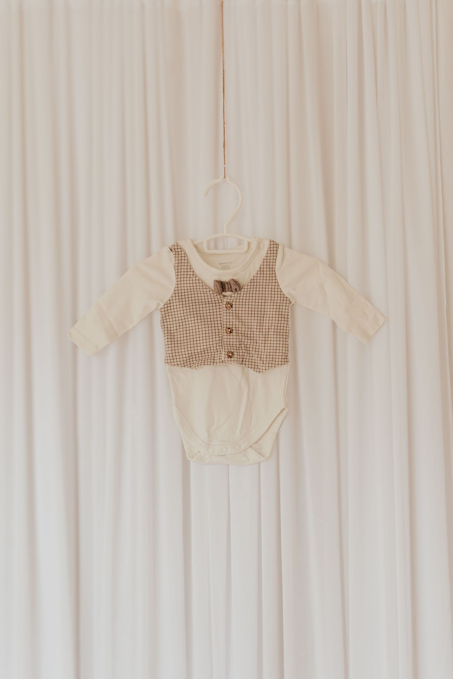 Ein Baby-Strampler mit cremefarbenem Stoff und braunem Gingham-Weste-Detail hängt an einem Kleiderbügel vor einem weißen Vorhang.
