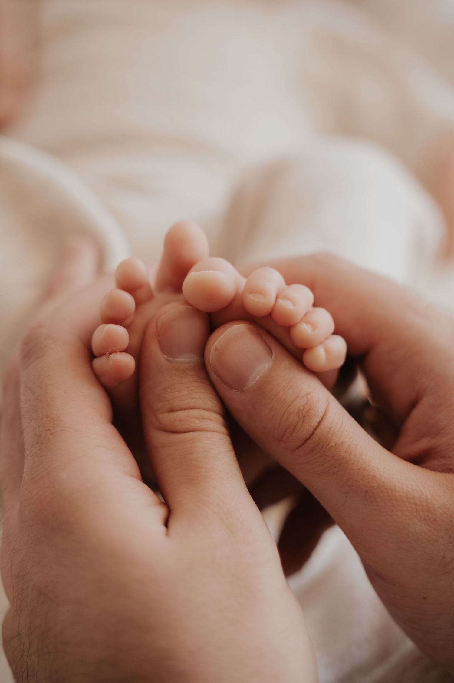 Adult hands gently cradling tiny newborn baby feet on a soft white blanket, close-up detail shot.